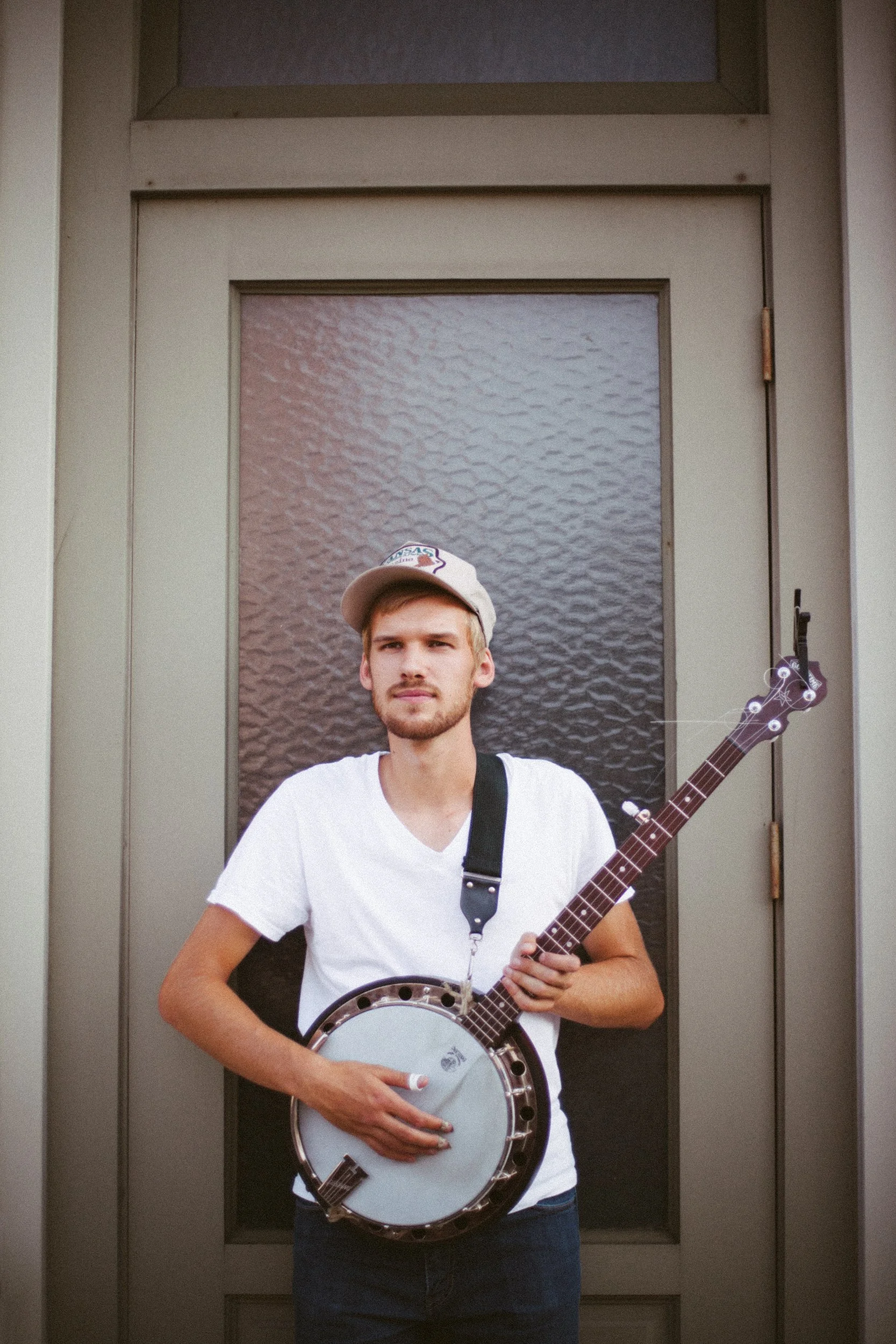 A young man with light brown hair, wearing a beige cap, white t-shirt, and black shorts, holding a banjo in front of a gray door with textured glass panels.