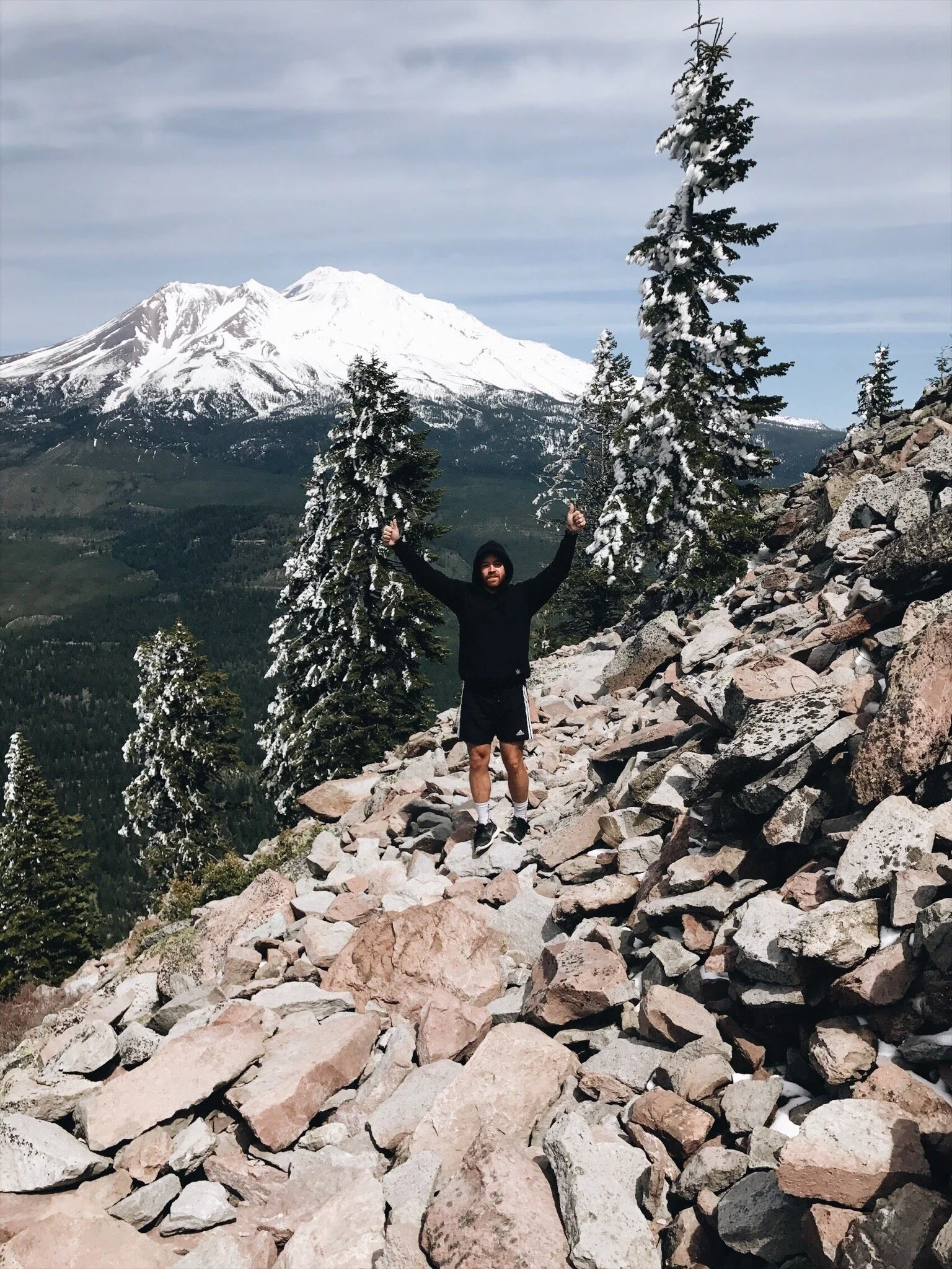 A person stands on a rocky mountain trail with snow-covered trees and a snow-capped mountain in the background, giving a thumbs-up gesture.