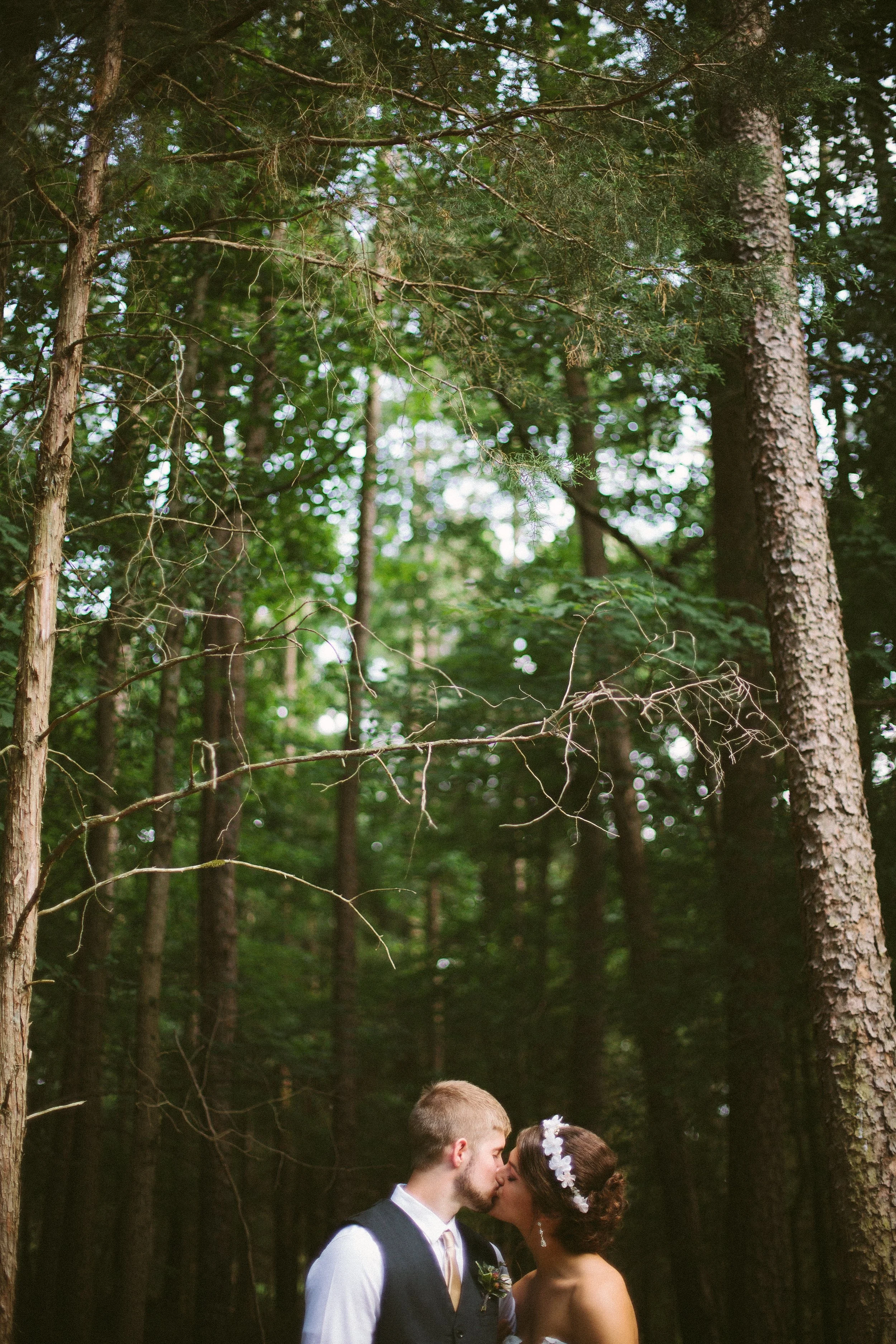 A bride and groom kissing in a forested area with tall trees and green foliage.
