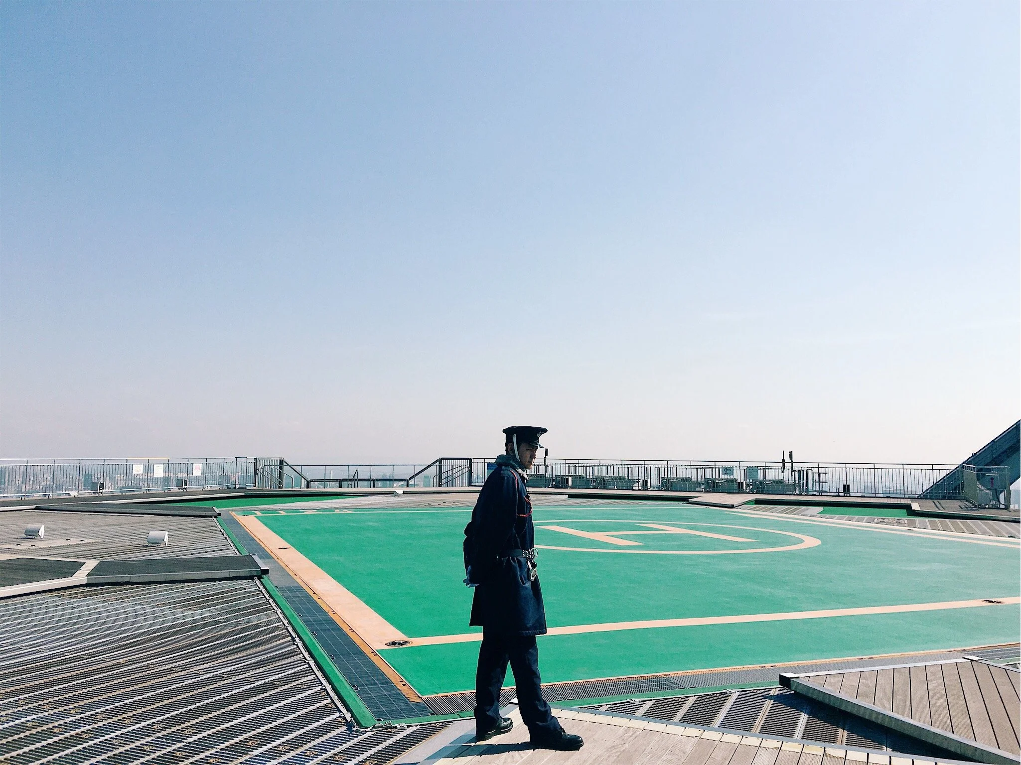 A person in a dark uniform and cap is standing on a rooftop helipad with green markings, surrounded by safety railings. The sky is clear with no clouds, and the cityscape is faintly visible in the distance.