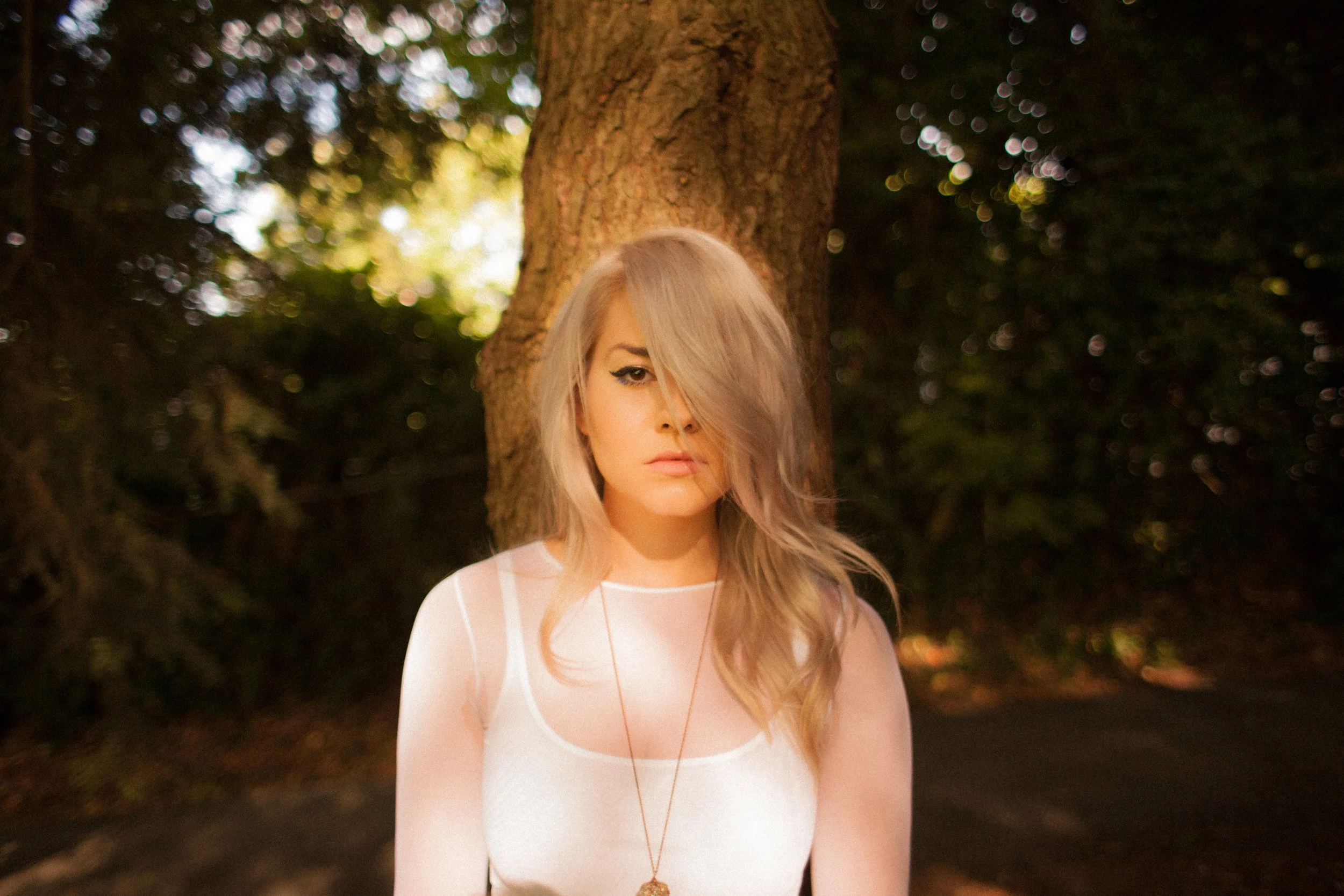 A woman with long blonde hair covering one eye, wearing a white top and a gold necklace, standing outdoors in front of a large tree with blurred leaves and sunlight in the background.
