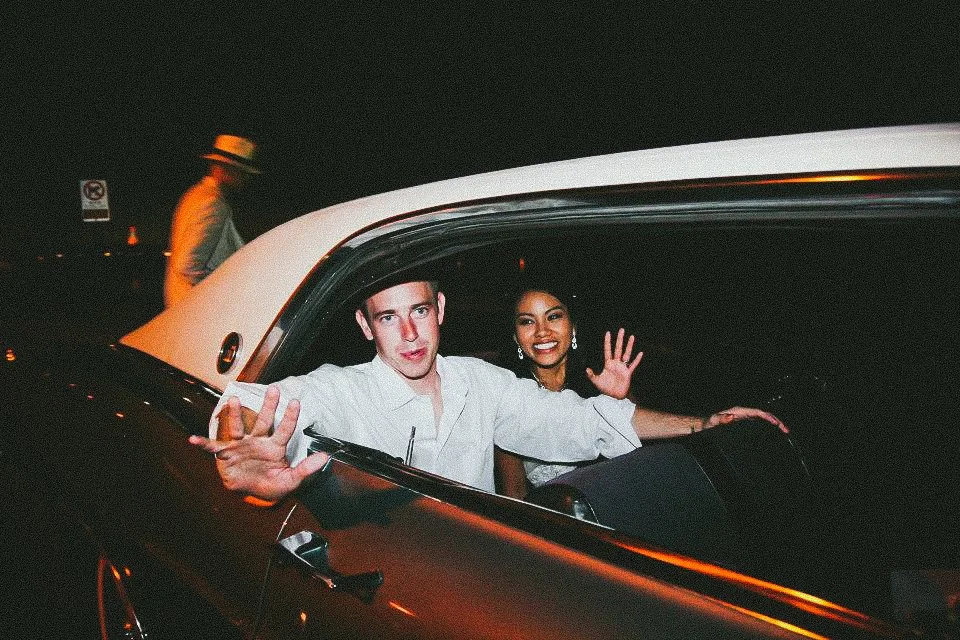 Two people sitting in a car at night, waving at the camera, with a third person walking in the background.