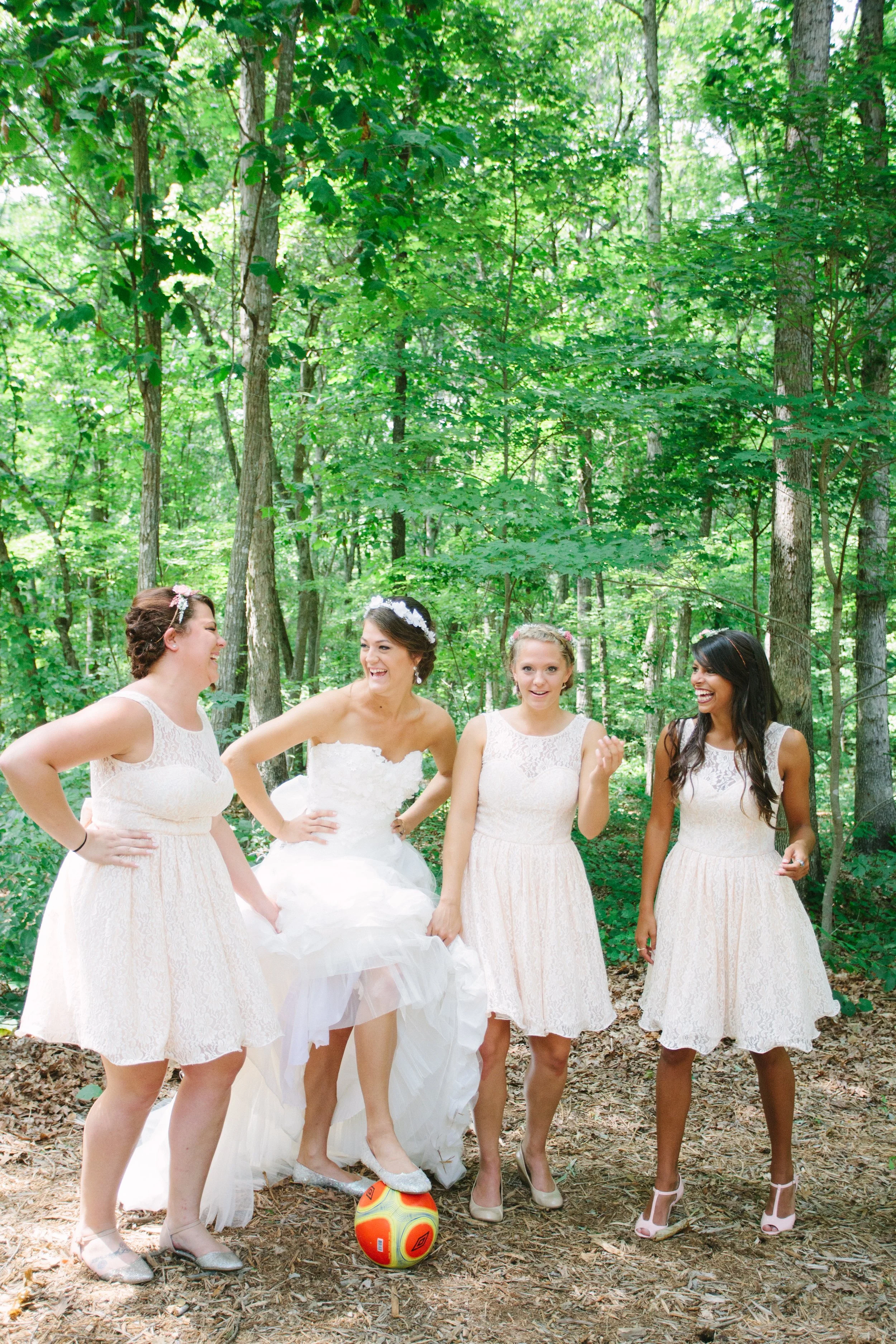 Group of women in wedding dresses playing with a soccer ball in a wooded forest area.