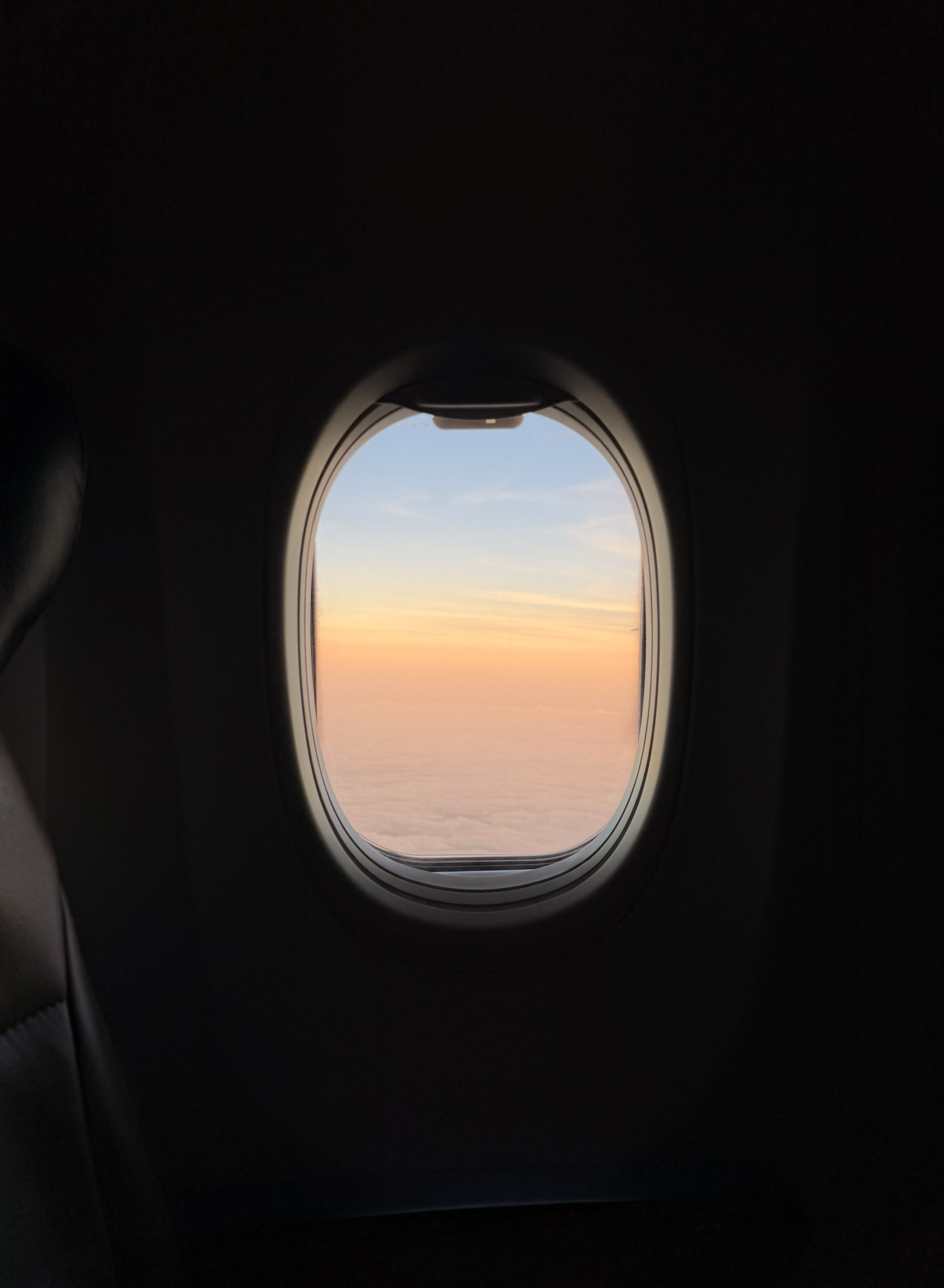 Photo taken from inside an airplane, showing an airplane window with a view of the sky during sunset or sunrise, with clouds below.