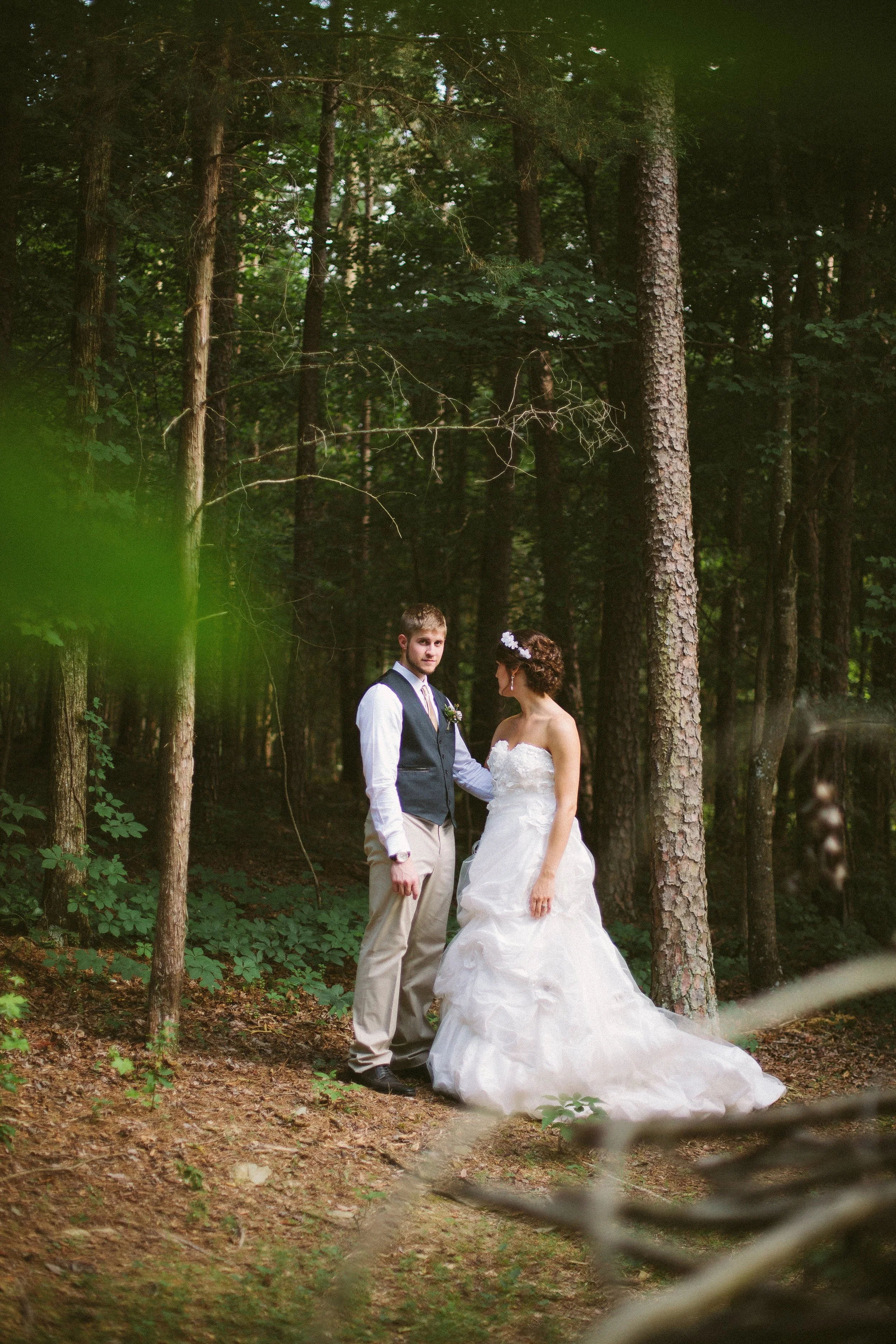 A couple dressed in wedding attire standing in a dense forest, surrounded by tall trees and green foliage.
