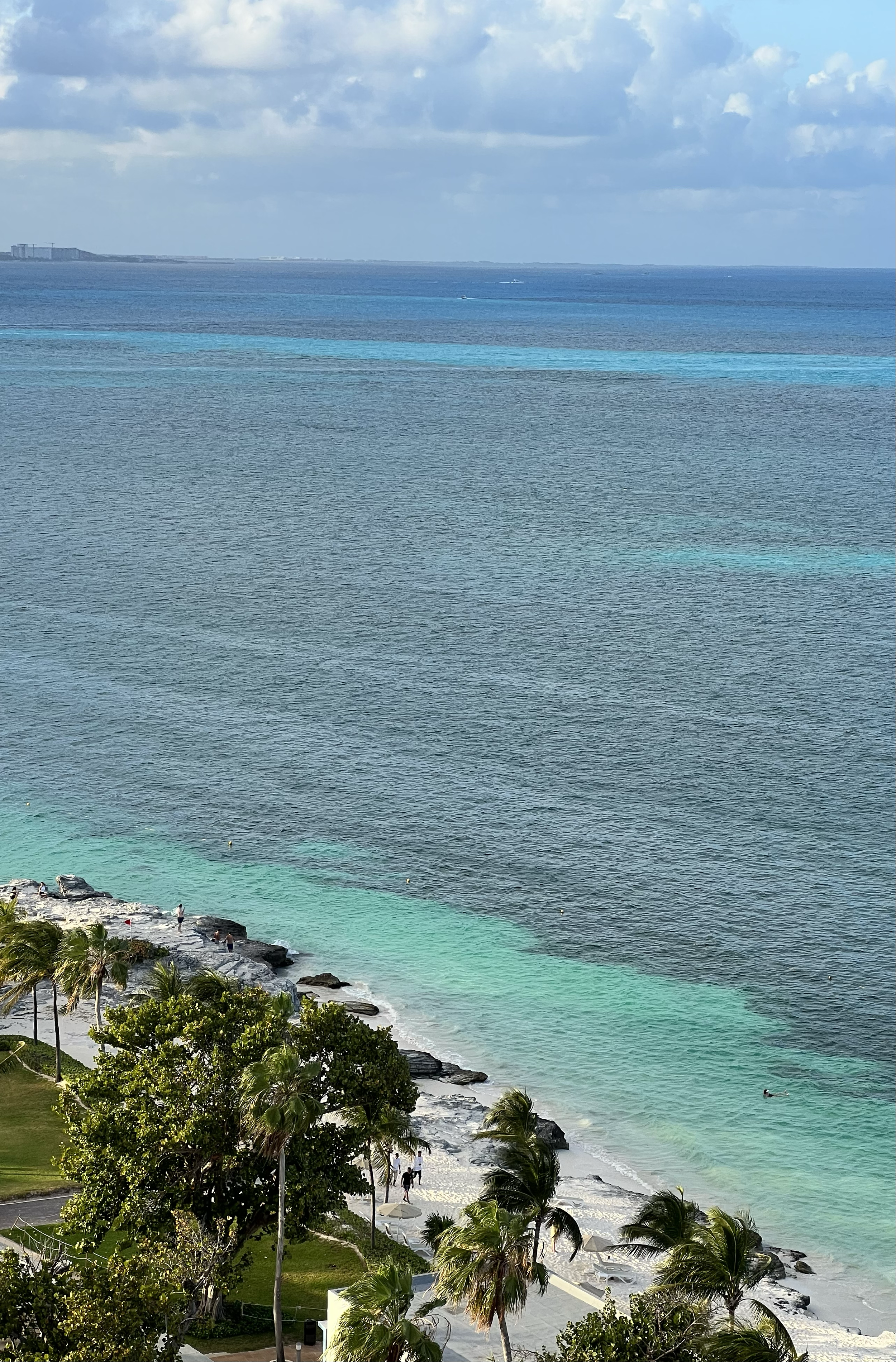 A scenic view of a beach with turquoise water, palm trees, and a blue sky with clouds. People are walking on the beach and relaxing under umbrellas, with a rocky shoreline in the foreground.