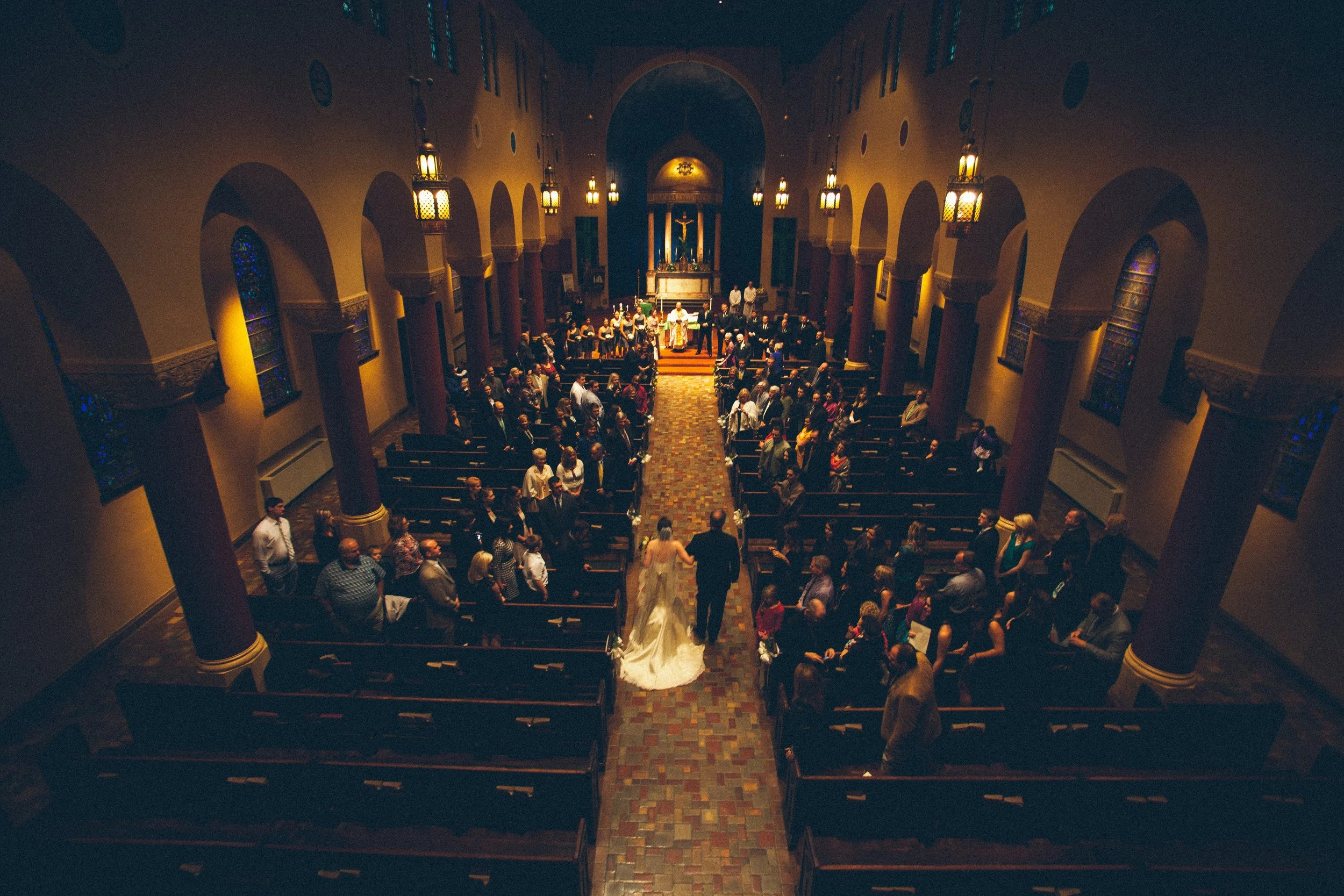 A church interior during a wedding ceremony with guests seated in pews and a bride and groom walking down the aisle.