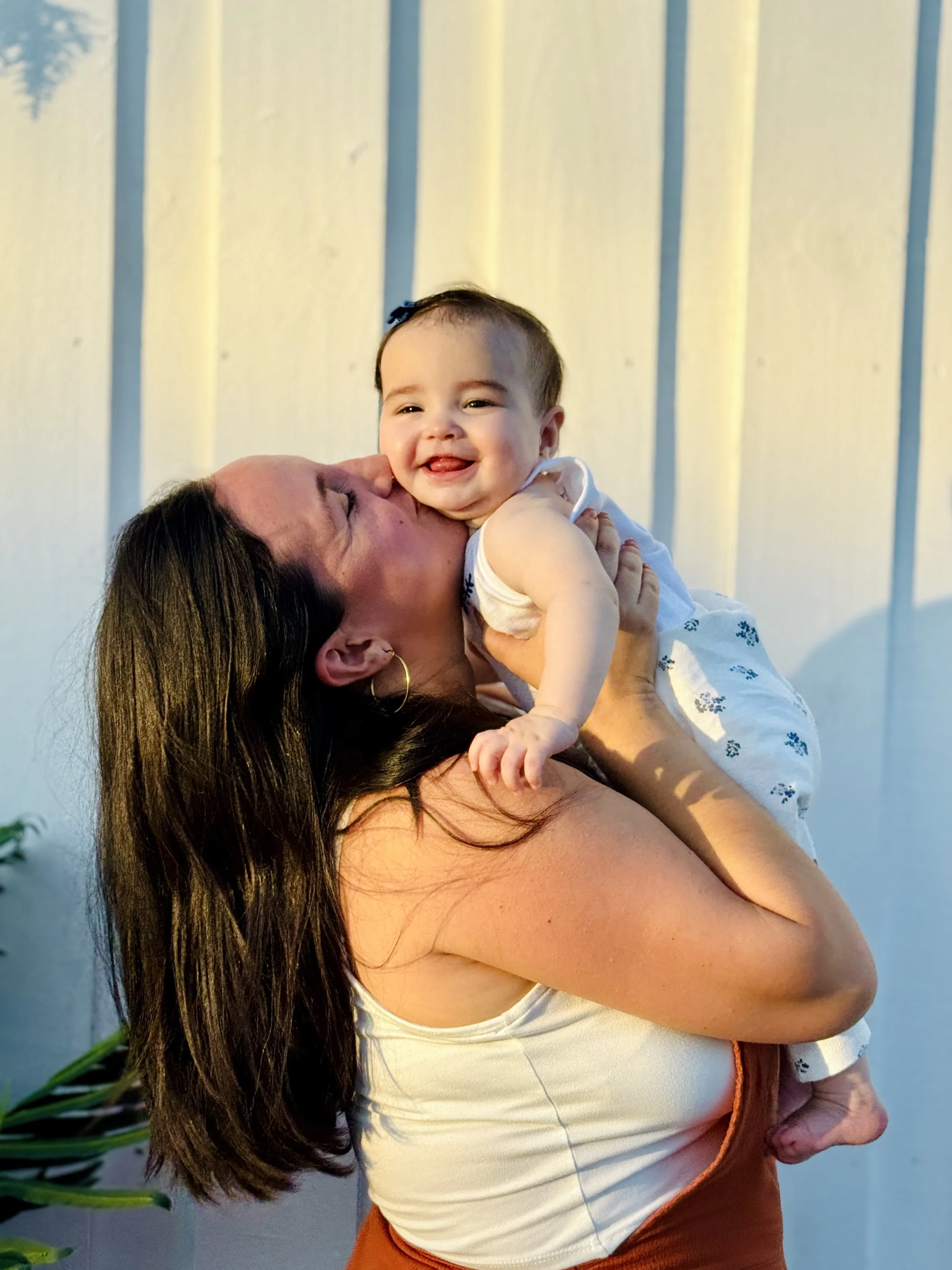 A woman with long brown hair holding a smiling baby girl with short curly hair, against a light-colored wall background.