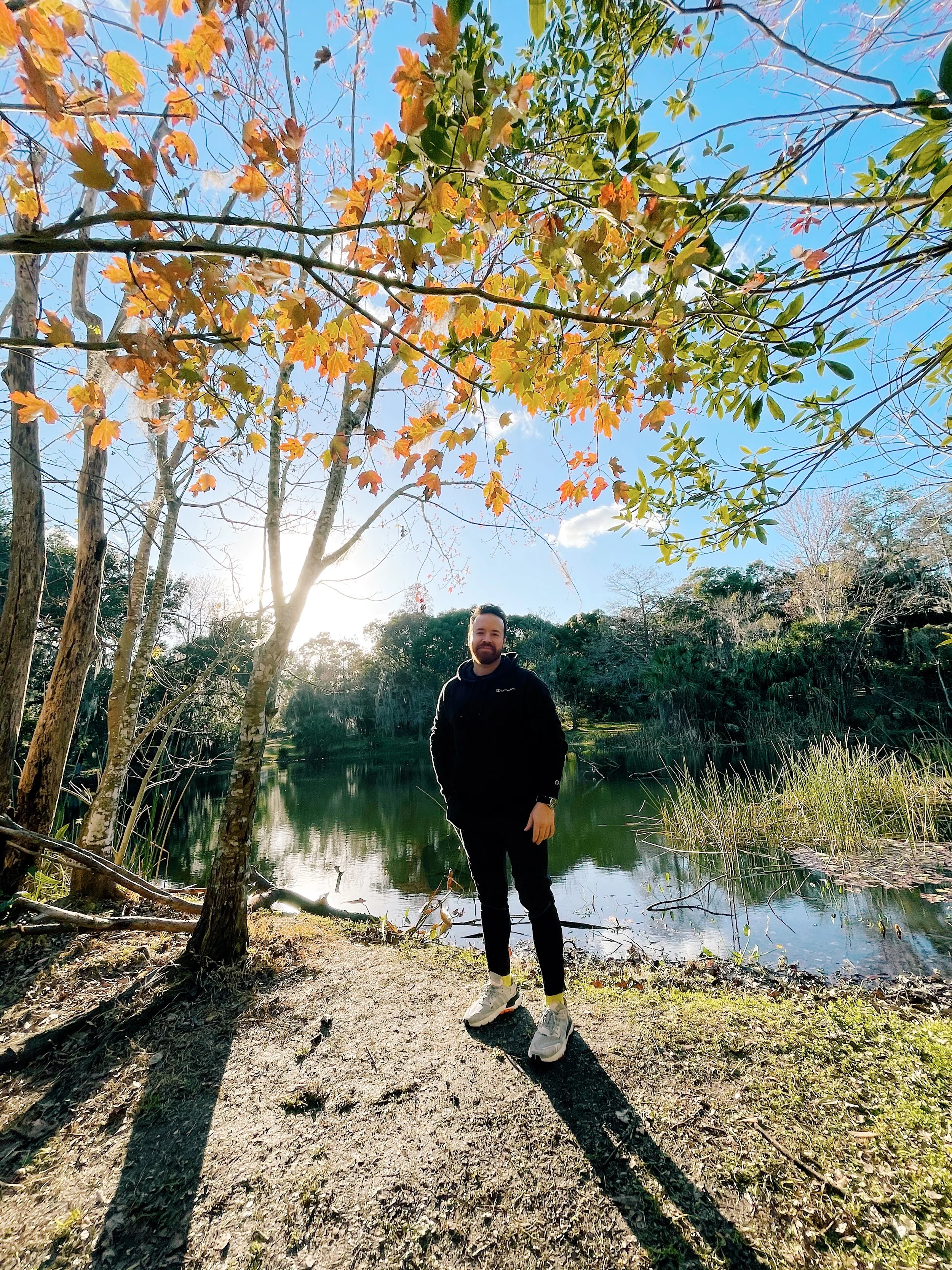A man with a beard and dark clothing standing by a lake with trees in autumn foliage, bright blue sky, and the sun shining in the background.