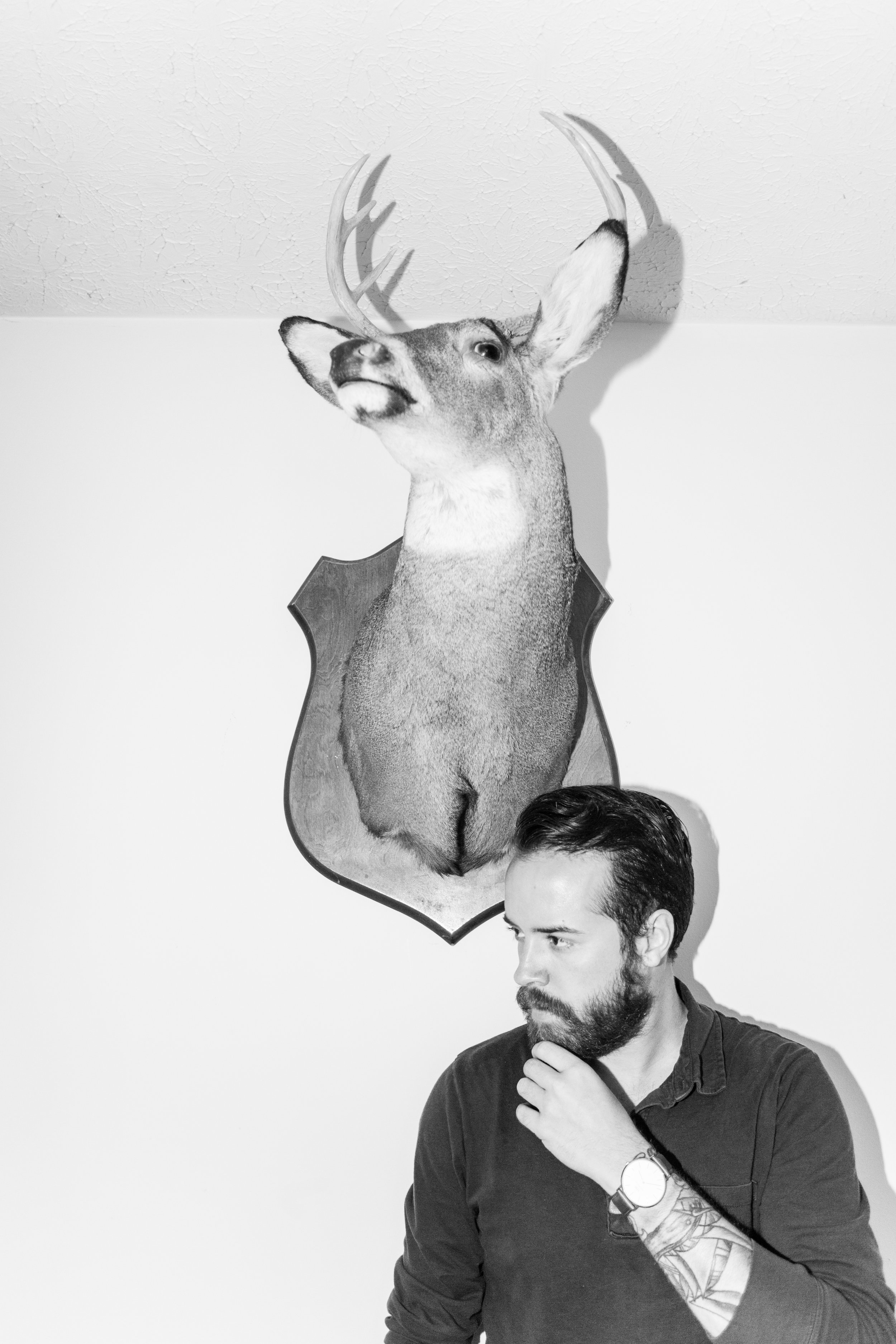 A bust of a deer head mounted on a wall, with a man's head and hand visible in the foreground. The man has a beard, tattoos on his arm, and is looking away thoughtfully.