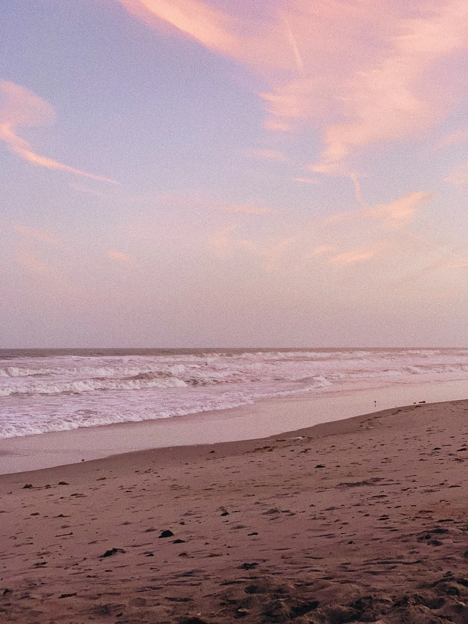 A beach at sunset with pink and purple clouds in the sky and gentle waves on the shoreline. Sand and small rocks are visible in the foreground.