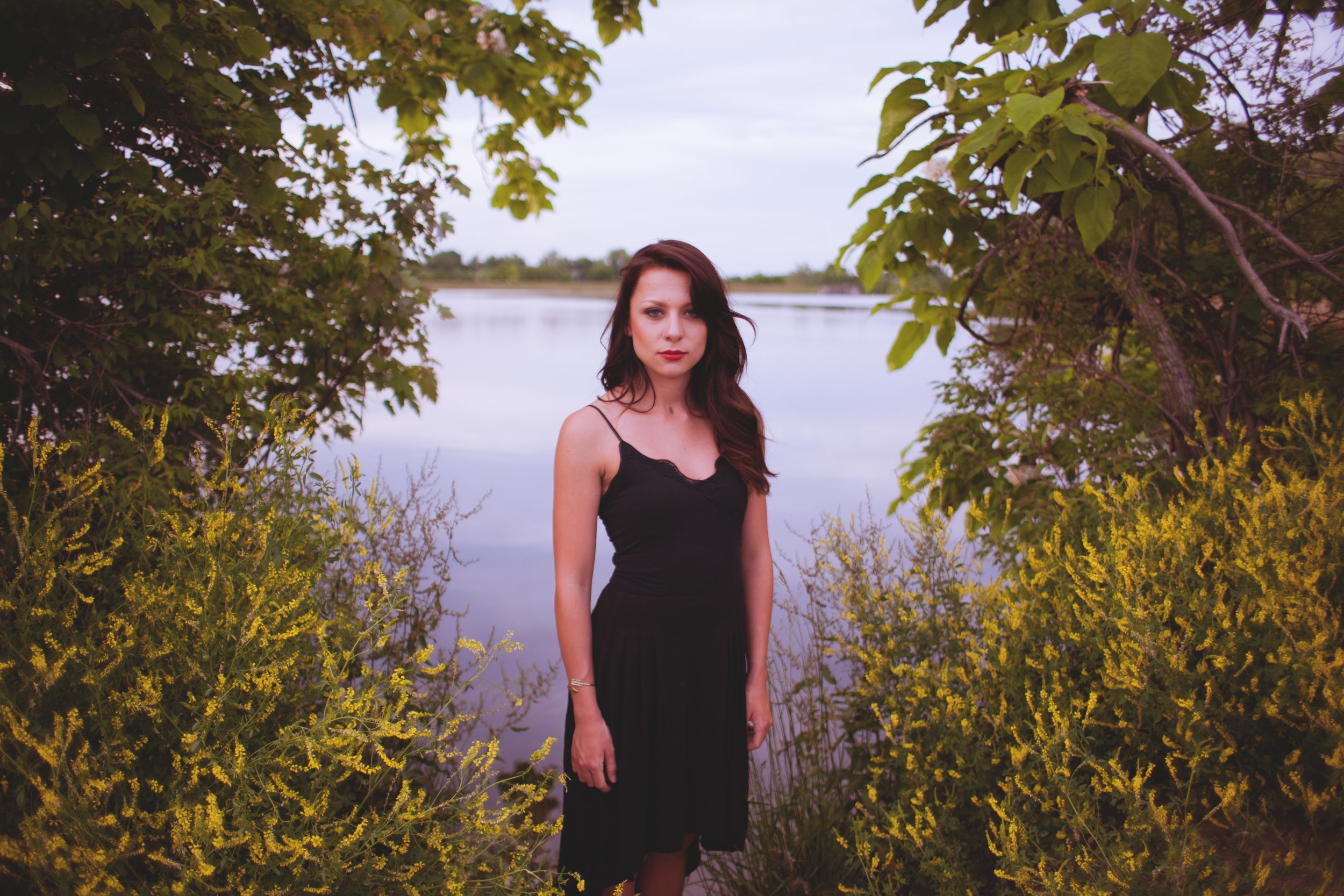 A woman in a black dress standing near a body of water, surrounded by green trees and yellow flowering bushes.
