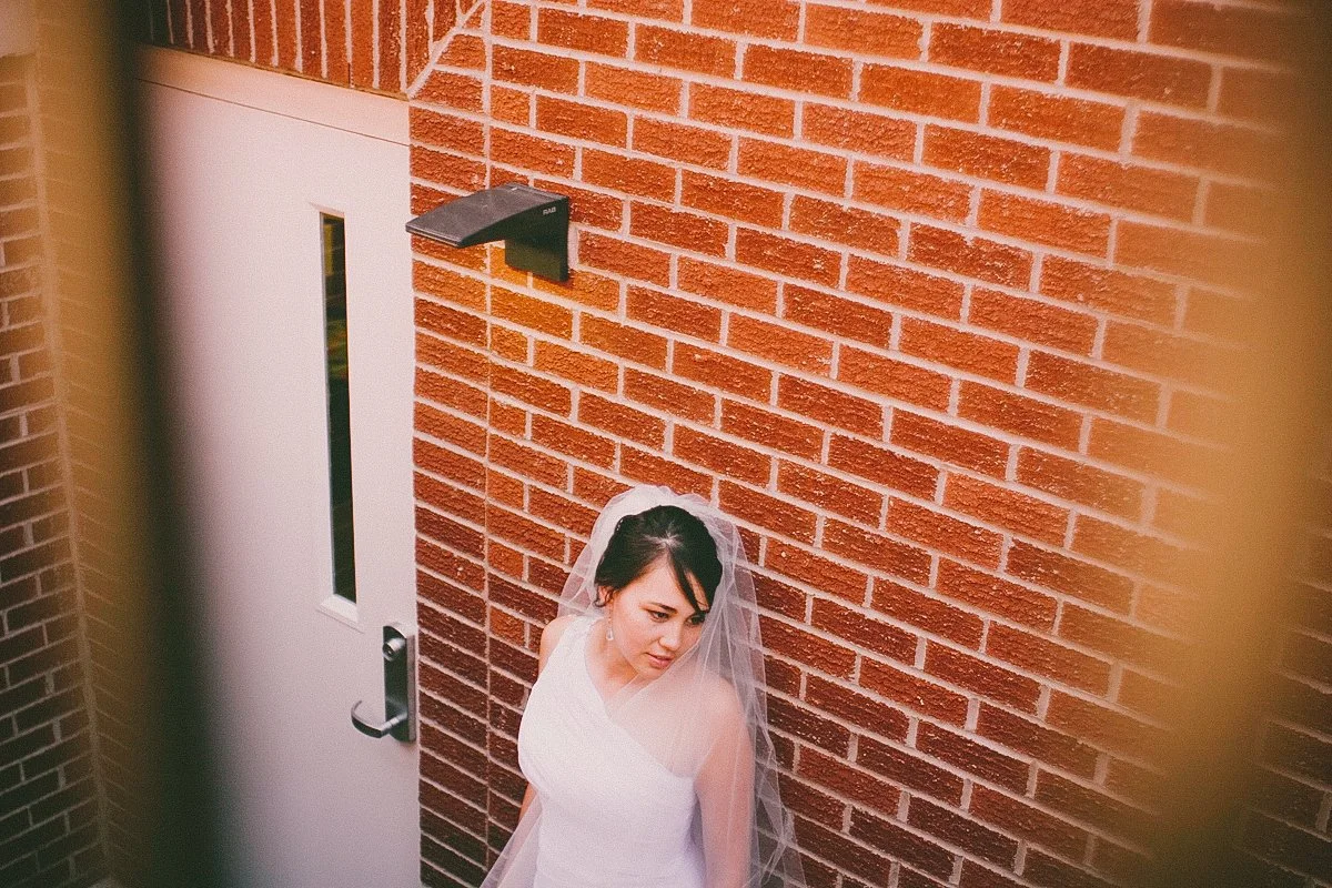 A bride in a white wedding dress with a veil poses outside near a red brick wall and a white door.