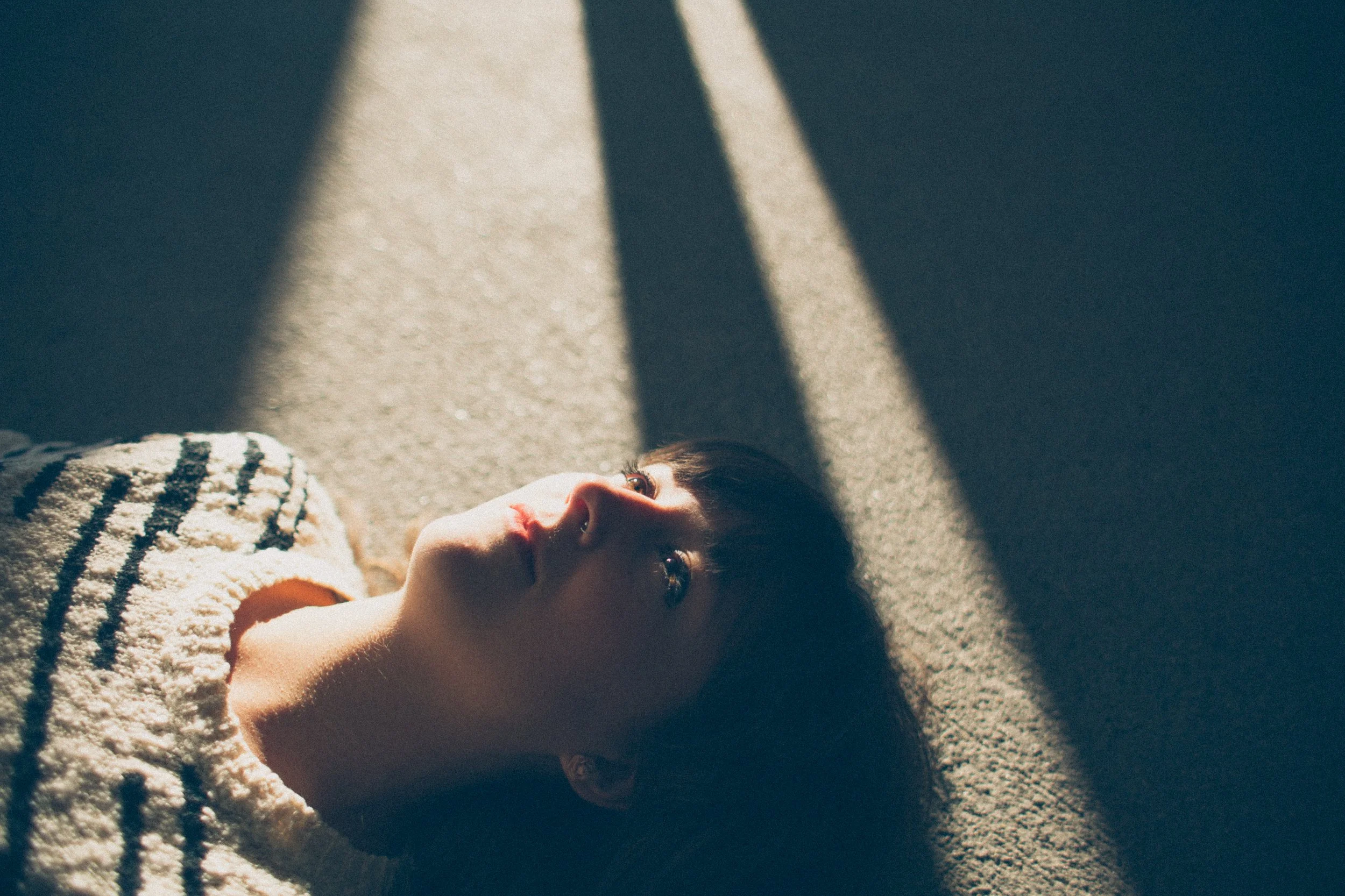 A young woman with dark hair and brown eyes lying on her back on the ground, looking up, with sunlight casting shadows.