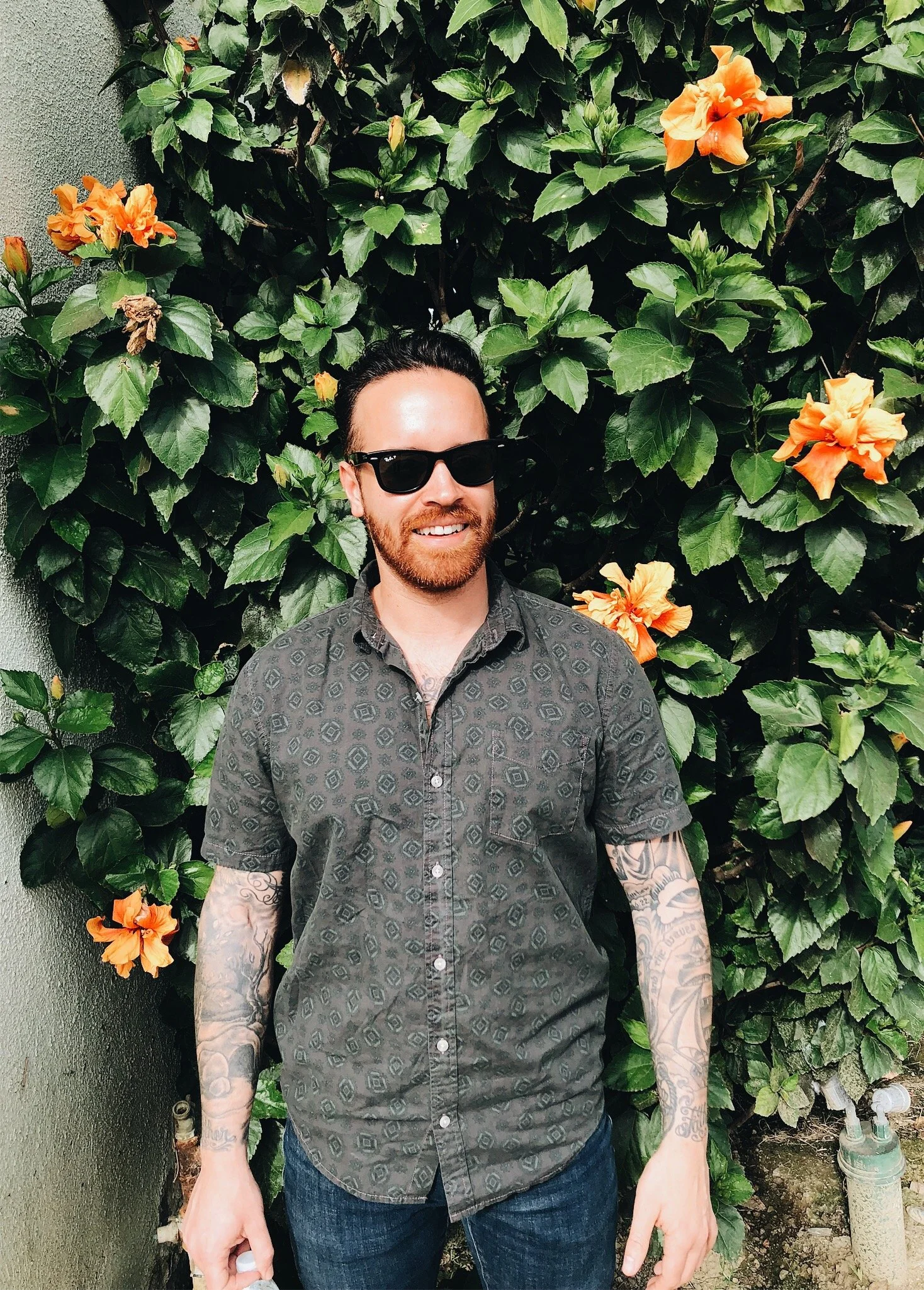 A man with sunglasses, a beard, and tattoos wearing a patterned shirt stands in front of green leaves and orange flowers.