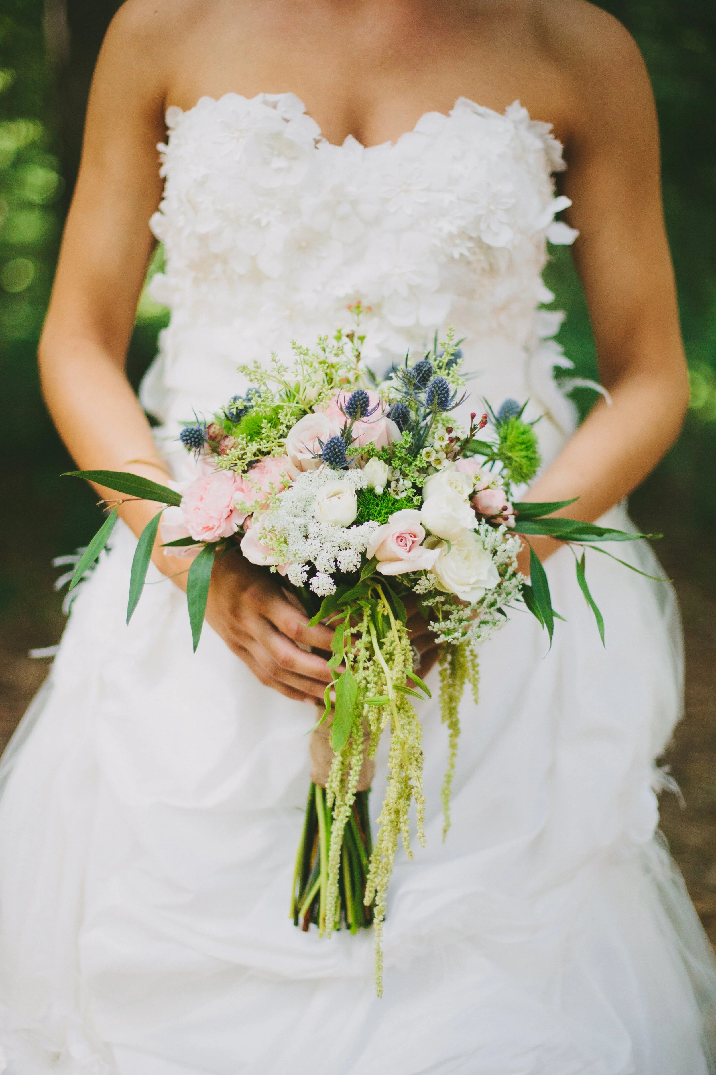 A woman in a white strapless wedding dress with floral embellishments, holding a bouquet of pink, white, and purple flowers with greenery, outdoors in a natural setting.