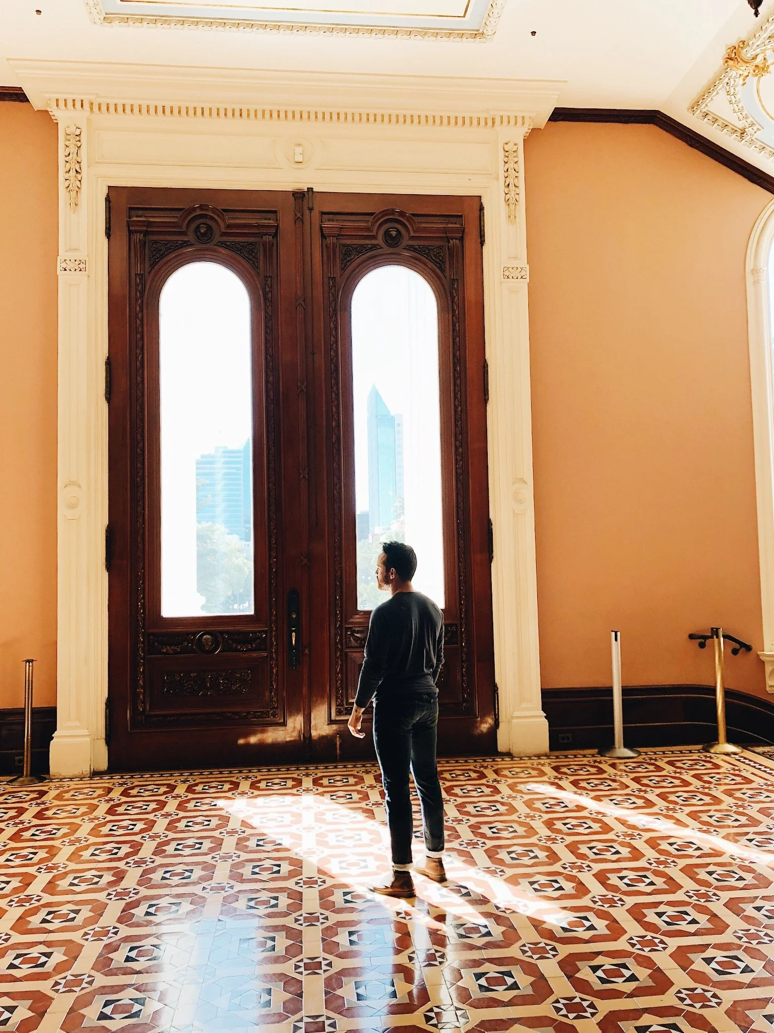 A man standing in front of ornate wooden double doors with tall windows, in a room with decorative patterned tile flooring, beige walls, and detailed crown molding. The backdrop shows city buildings through the windows.
