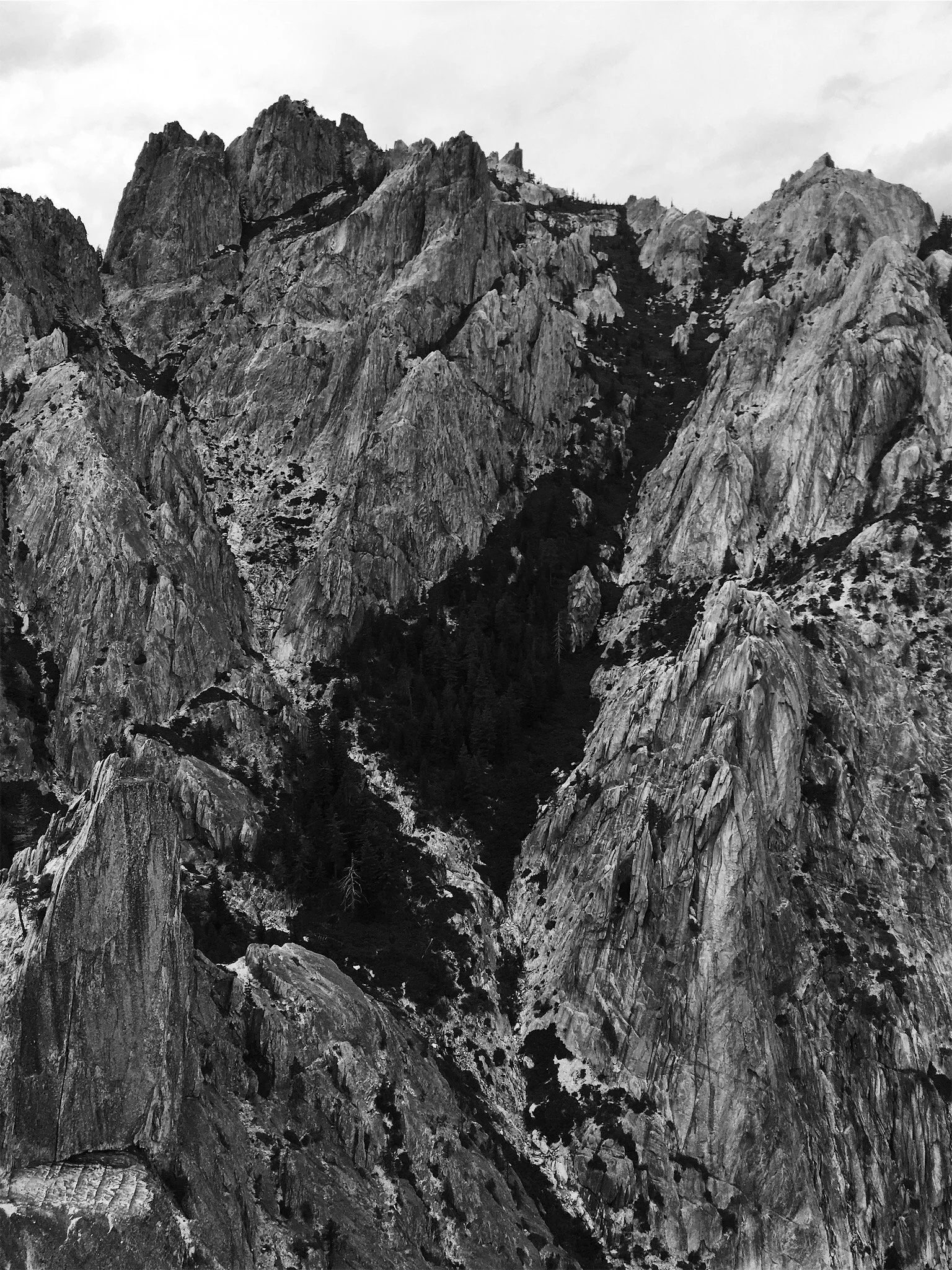 A black and white photograph of rugged mountain peaks with steep rocky slopes and some trees at the base.
