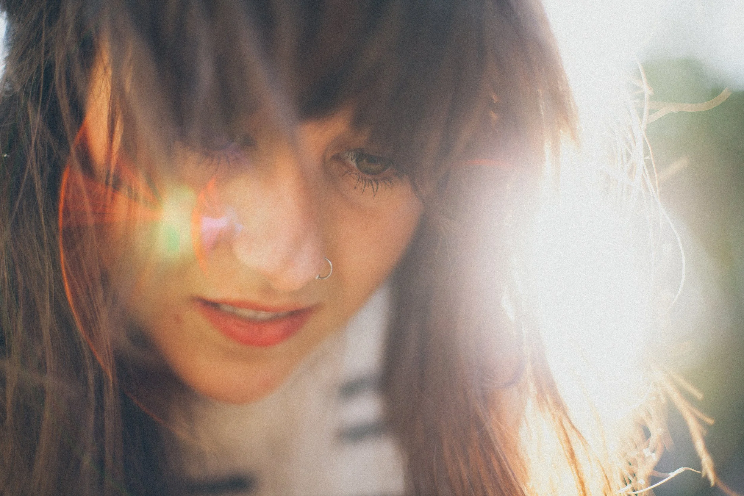 Close-up of a woman with long auburn hair, looking down with a slight smile, outdoors in natural light.