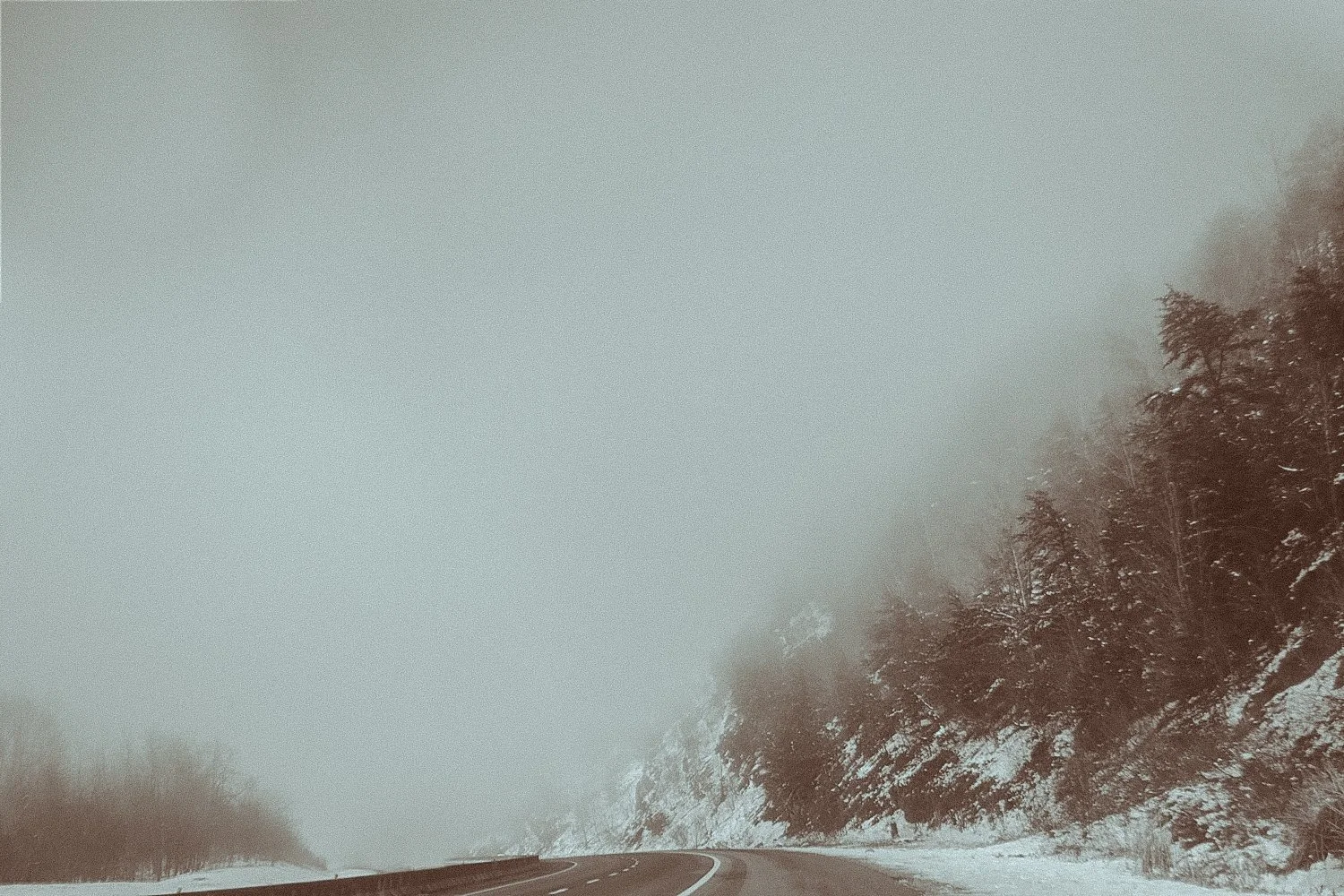 Snow-covered mountain road with foggy skies and trees on the side.