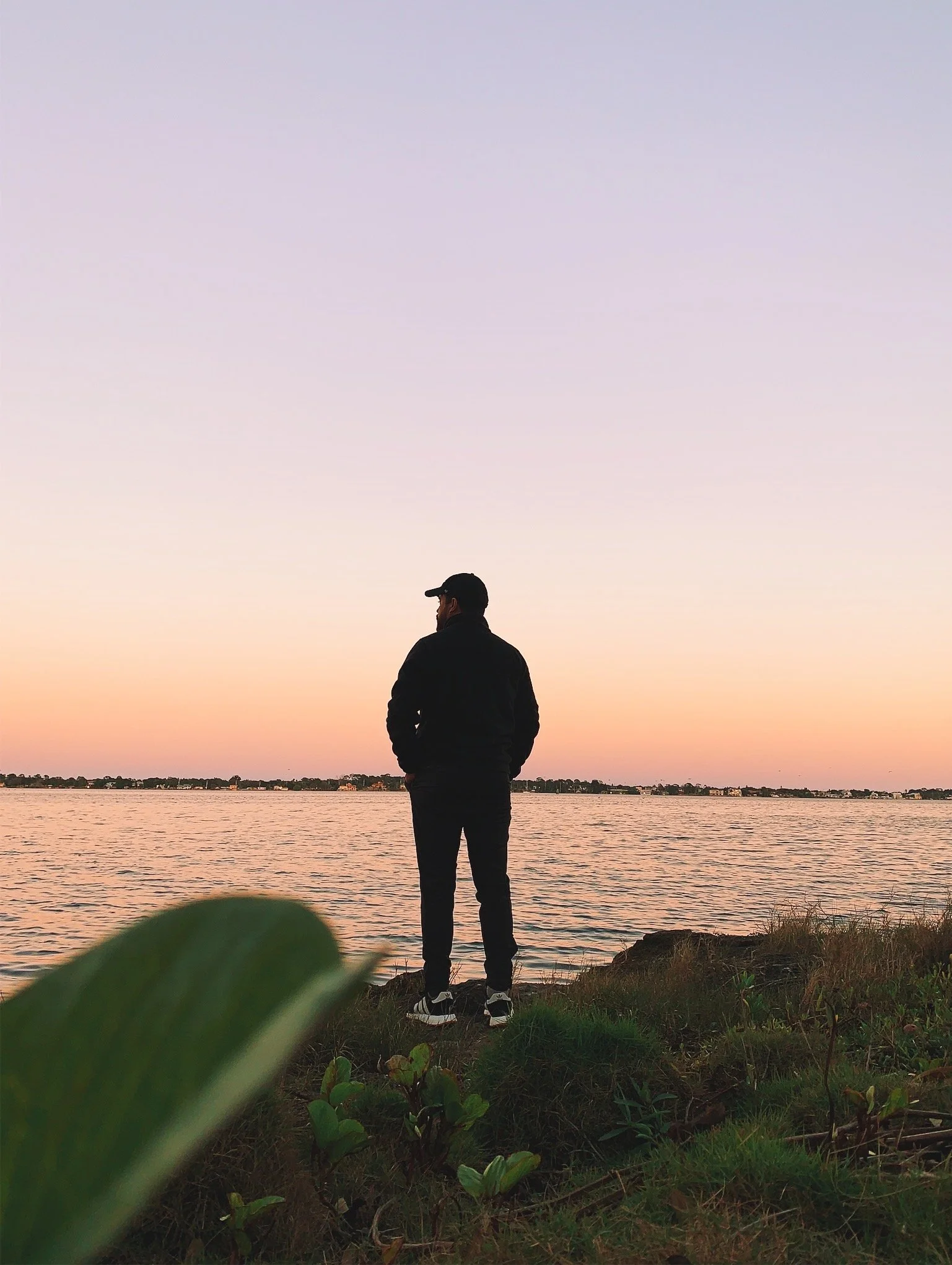 A person stands on the grassy shore of a body of water during sunset, with a clear sky fading from pink to purple, and a distant shoreline visible across the water.