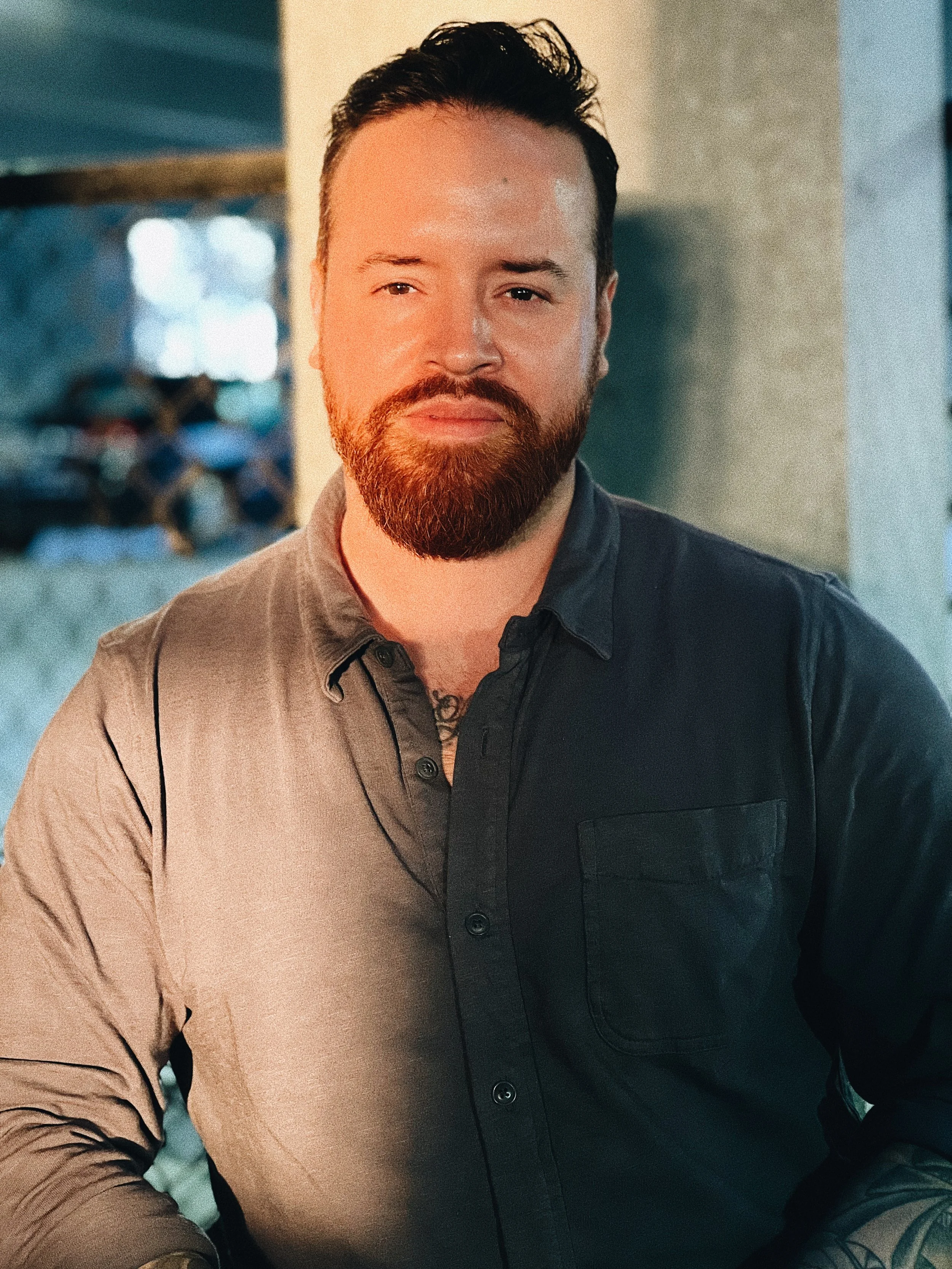 Close-up of a bearded man with dark hair, wearing a black shirt, standing indoors with a blurred background.