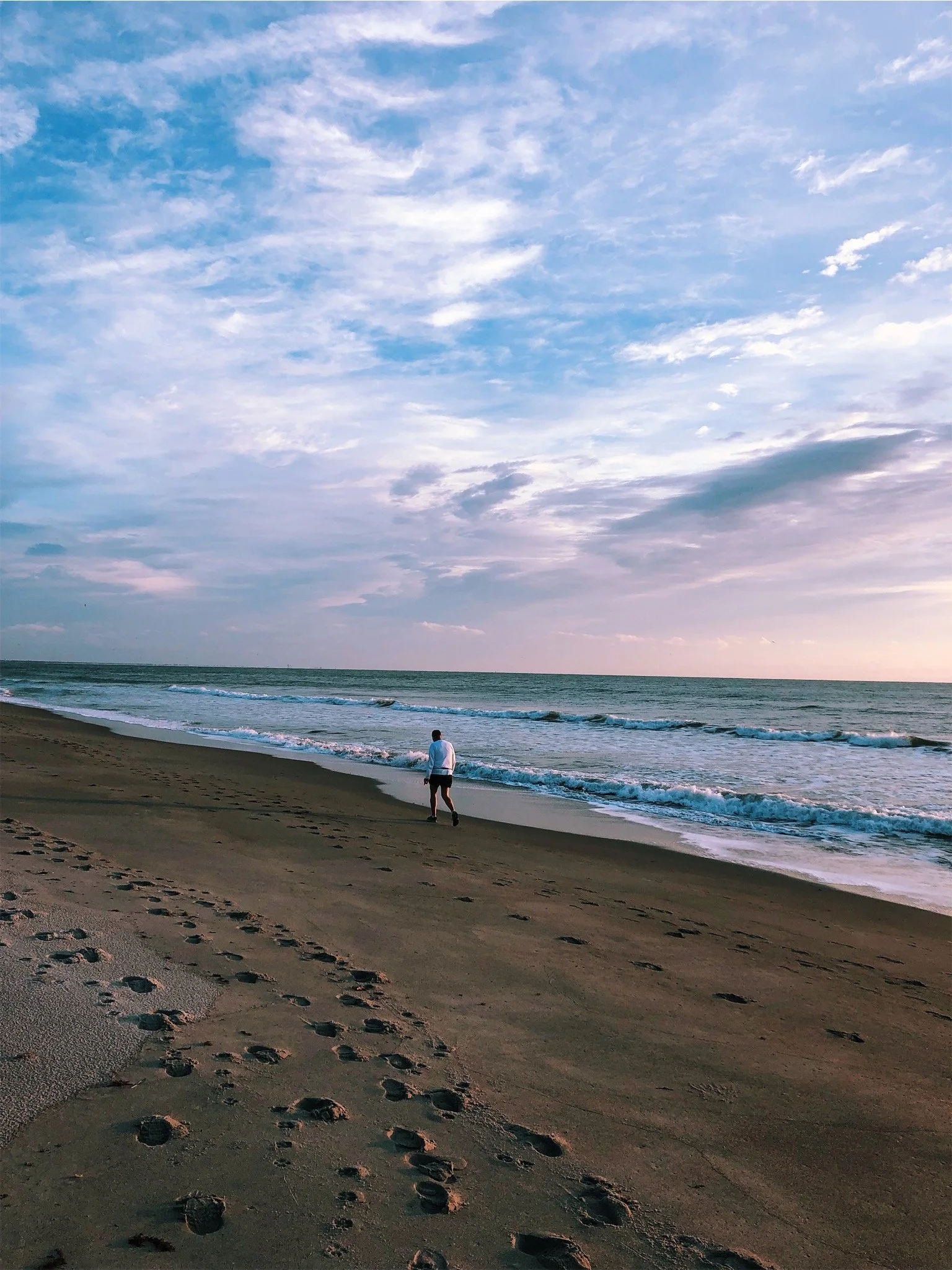A person walking alone on a sandy beach near the ocean during sunset or sunrise, with footprints in the sand and a sky with scattered clouds.