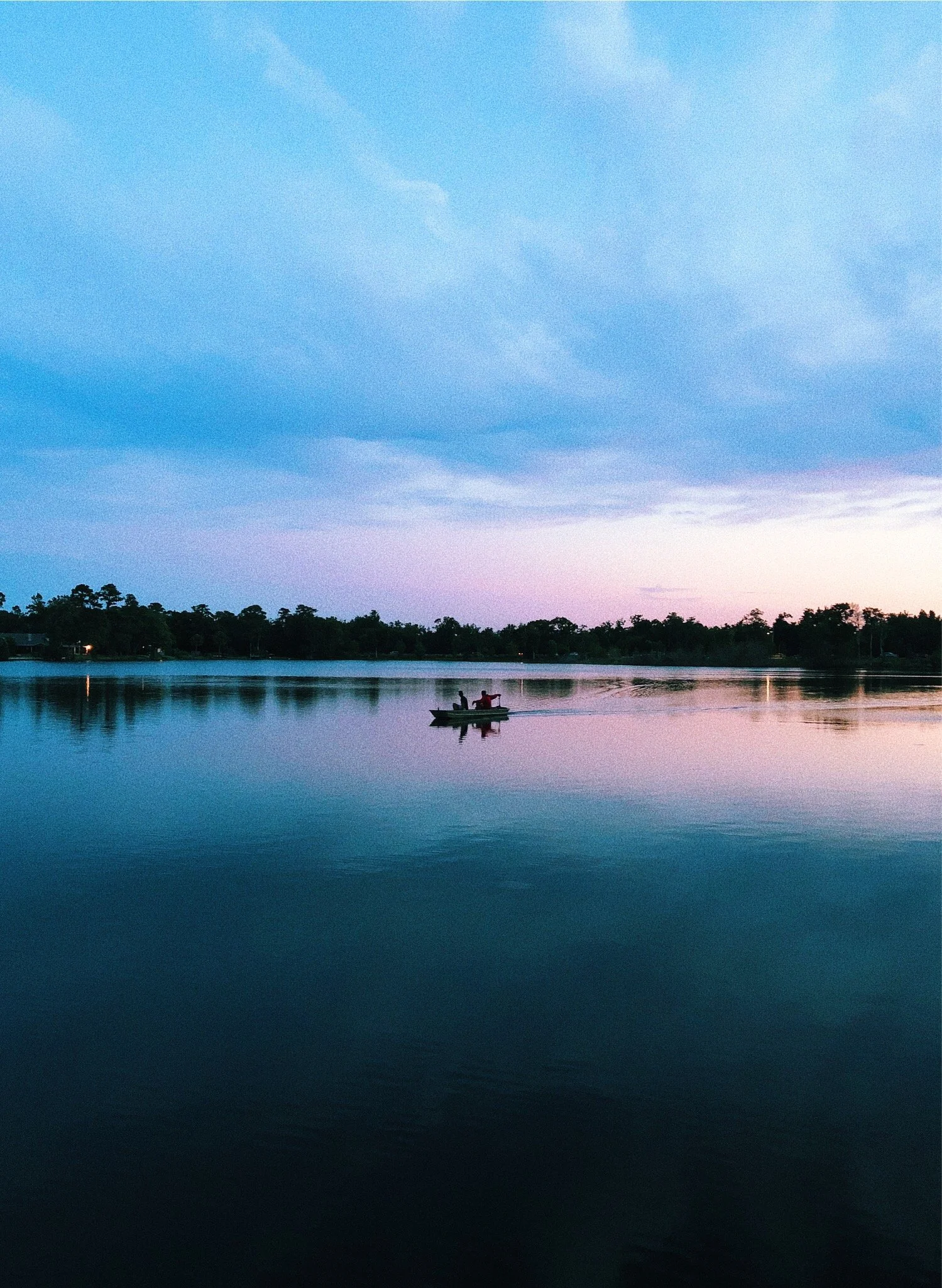 A serene lake at dusk with a small boat carrying two people, calm water reflecting the sky's gradient from pink to blue, and a tree-lined shoreline in the background.