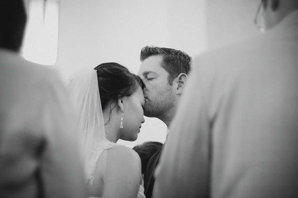 A black and white photo of a wedding ceremony showing a couple kissing, with the bride wearing a veil and earrings, and the groom with short hair and a beard, surrounded by guests.