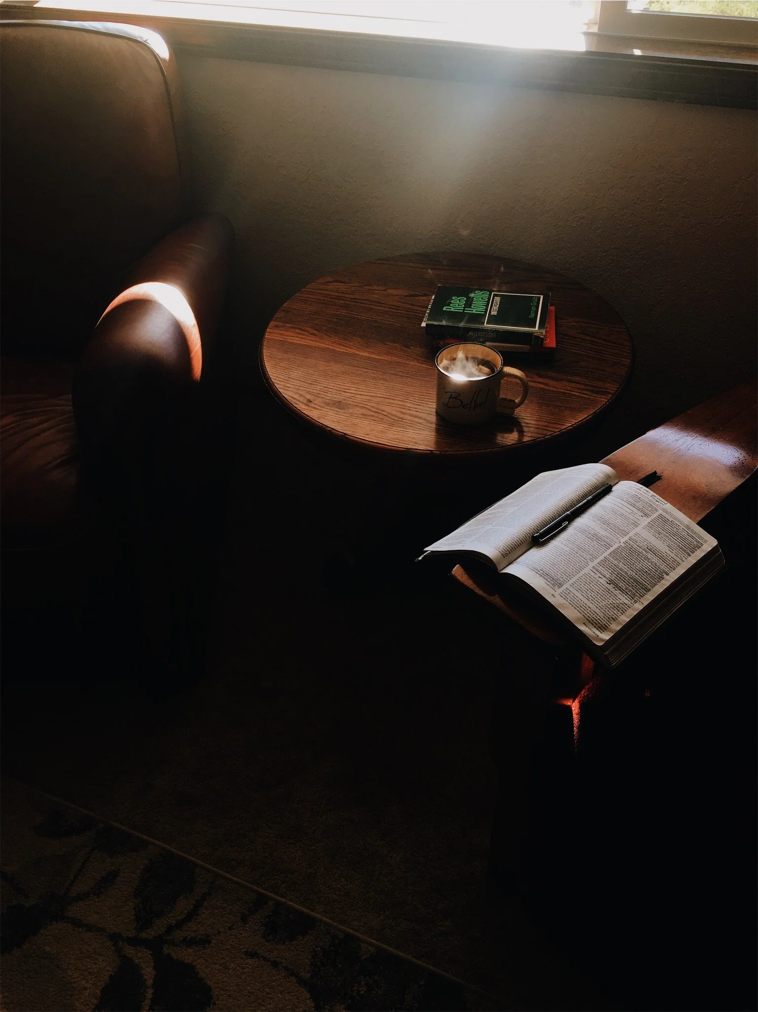 A cozy reading nook with a wooden table, a steaming coffee mug, an open book, and two closed books, with sunlight streaming through a window.