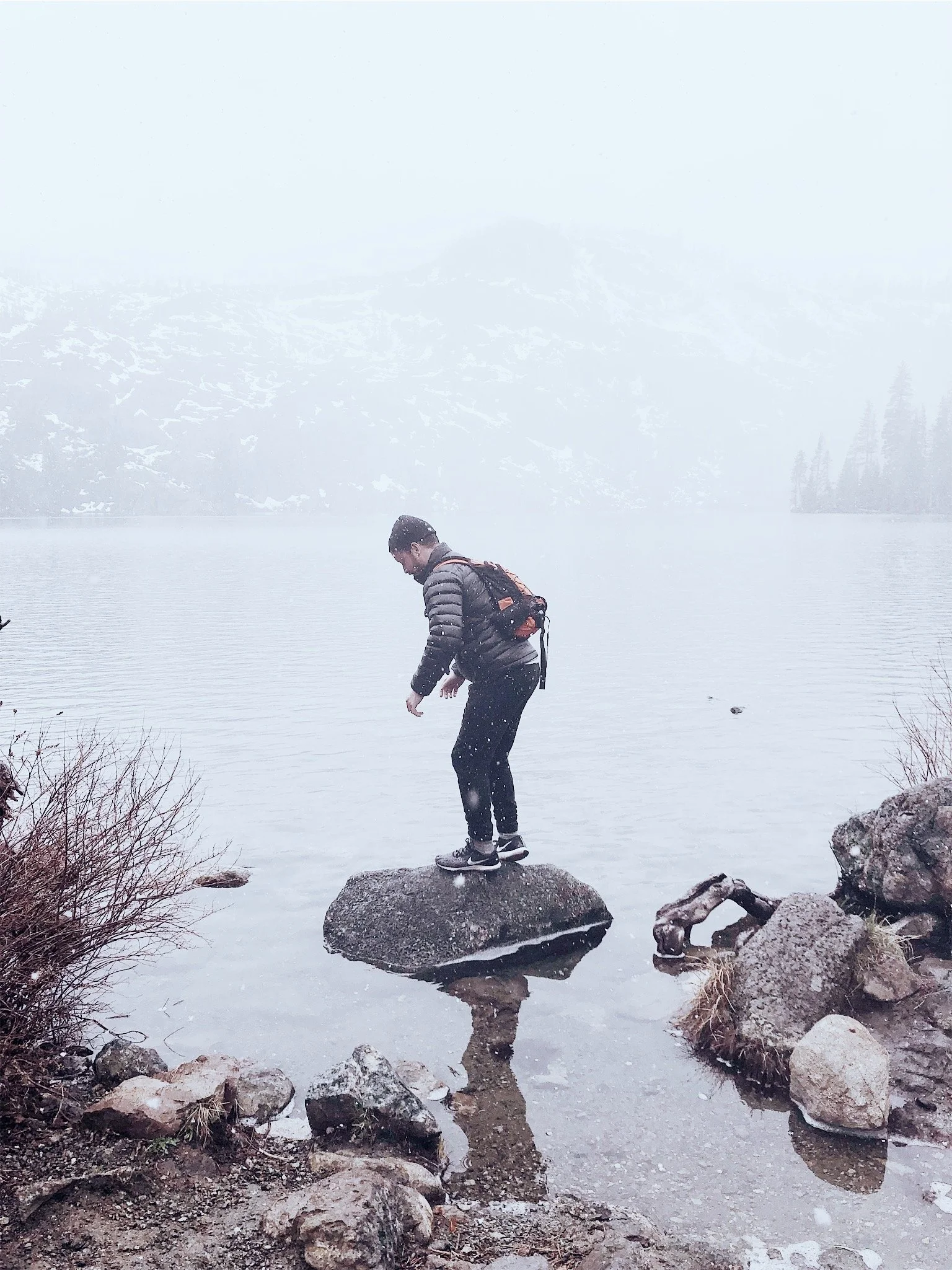 A person in a jacket and backpack standing on a rock by a lake in a foggy, snowy mountain landscape.