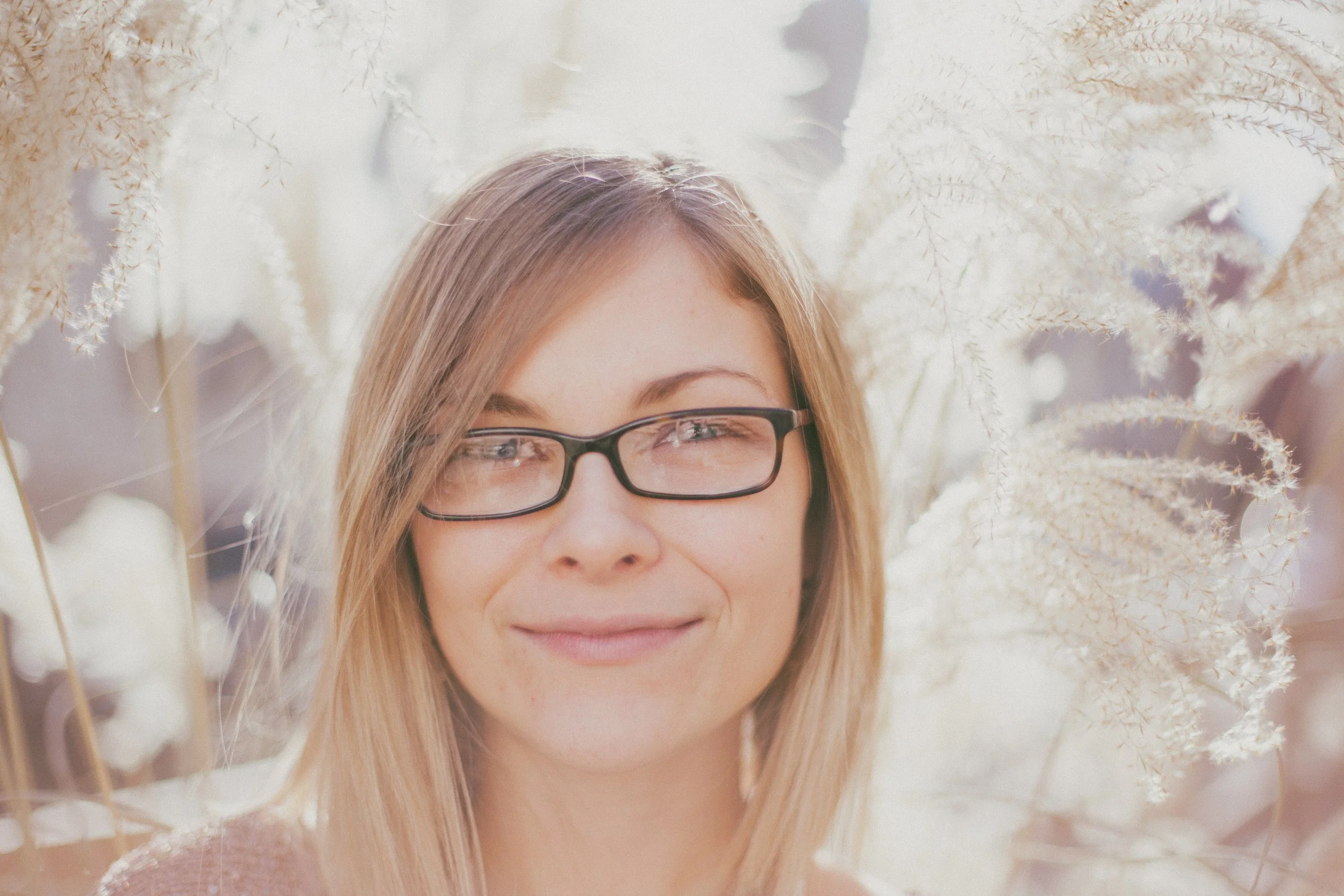 A young woman with blonde hair and glasses smiling, surrounded by white fluffy flowers.
