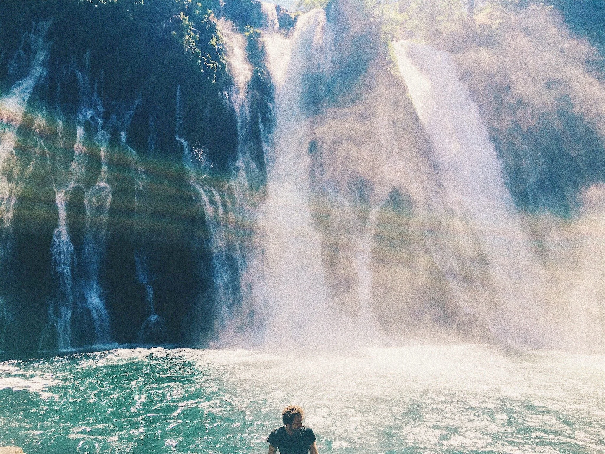 A person standing at the edge of the ocean, looking at a large waterfall in the distance with flowing water and spray.