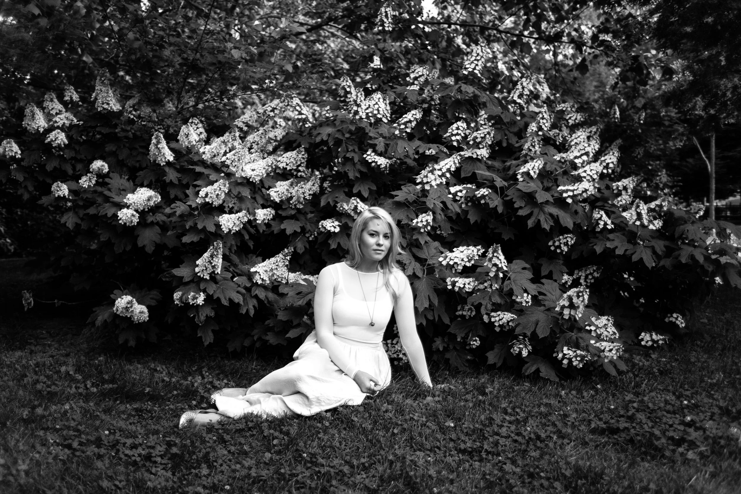 A woman sitting on the grass in front of large leafy plants with white flowers, in a park-like setting, black and white photograph.