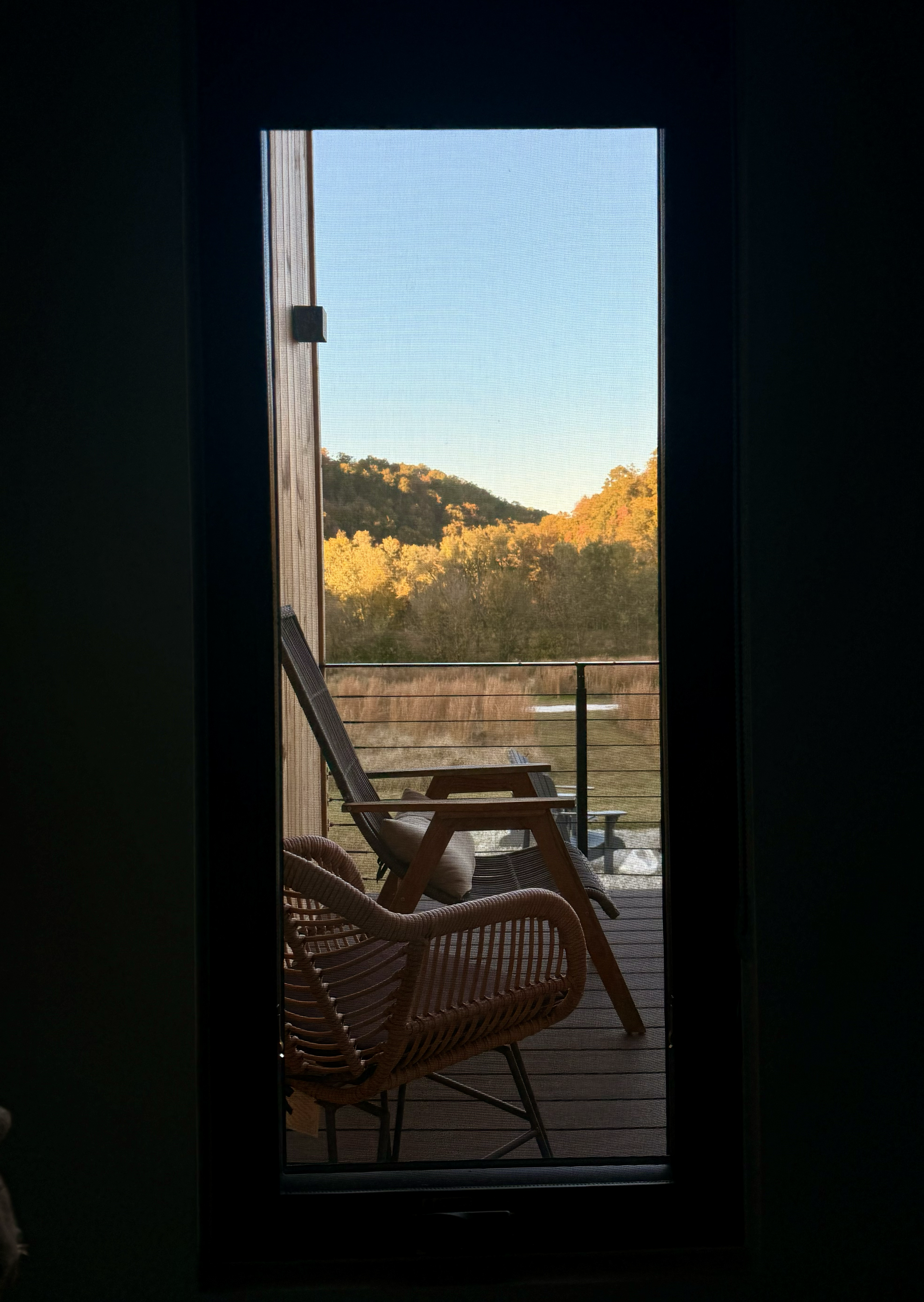 View through a door screen showing a balcony with wicker chairs, overlooking trees and hills with autumn foliage under a clear blue sky.