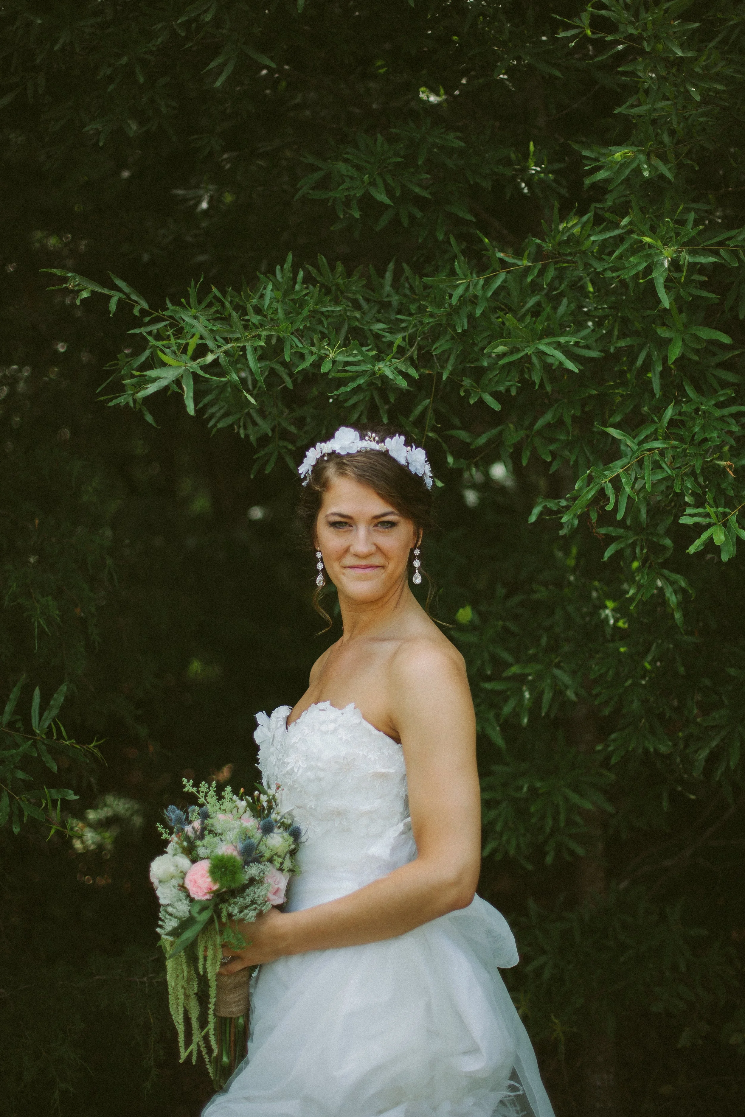 A bride standing outdoors in front of green leafy trees, holding a bouquet of flowers, wearing a white strapless wedding gown with floral details, and a floral headband, smiling softly.