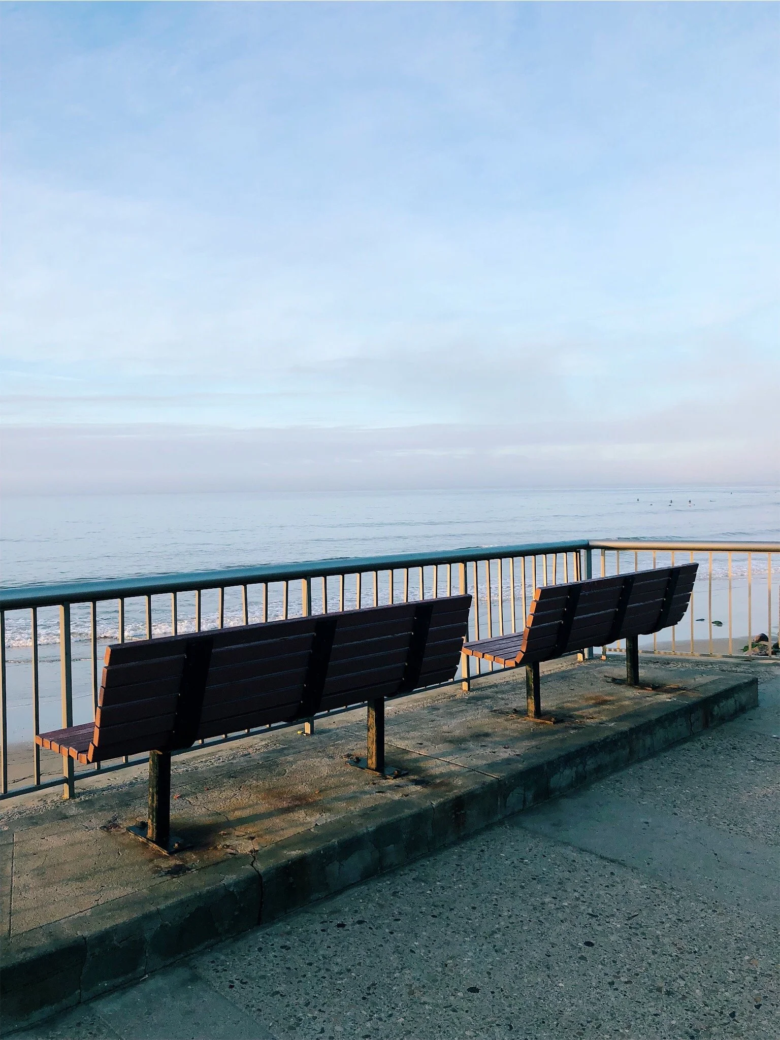 Three empty benches facing the ocean along a seaside promenade with a metal railing and cracked pavement, under a cloudy sky.