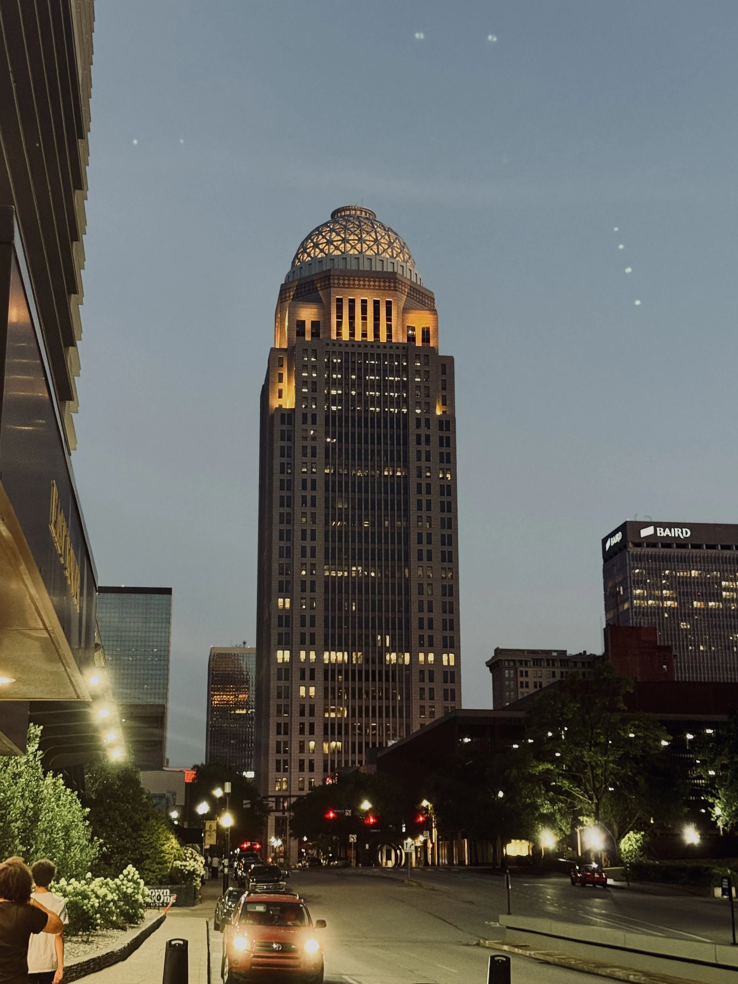 A cityscape at dusk featuring a tall skyscraper with a dome-shaped top illuminated with warm lighting. The street level shows parked cars, people walking, and greenery along the sidewalk, with a partly cloudy sky overhead.