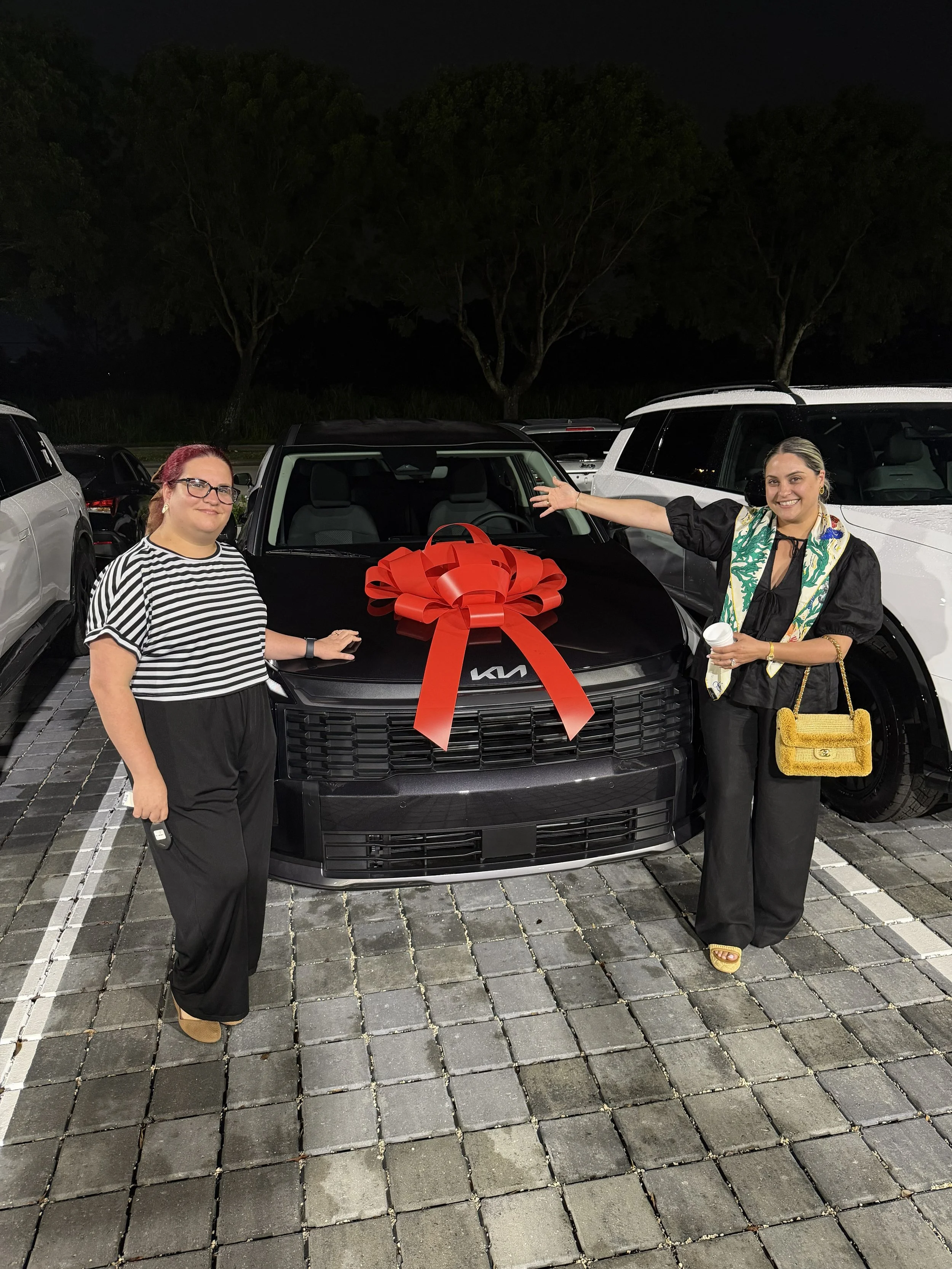 Two women standing next to a black Kia SUV with a large red bow on the hood, at night in a parking lot with trees in the background.