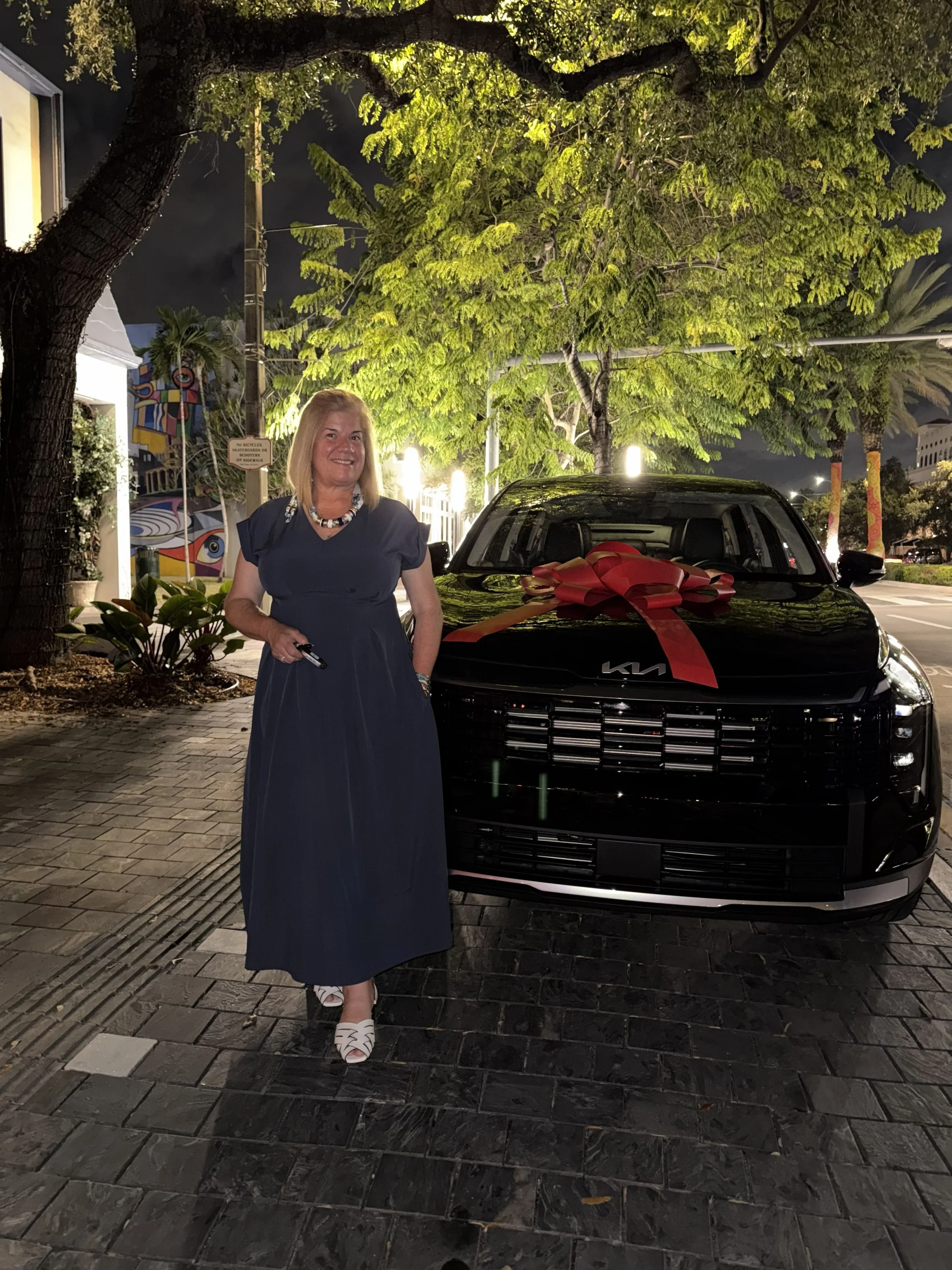 A woman in a blue dress with white shoes and a necklace is standing next to a black Kia car with a red ribbon on the hood. The scene is outdoors at night, with trees and streetlights in the background.