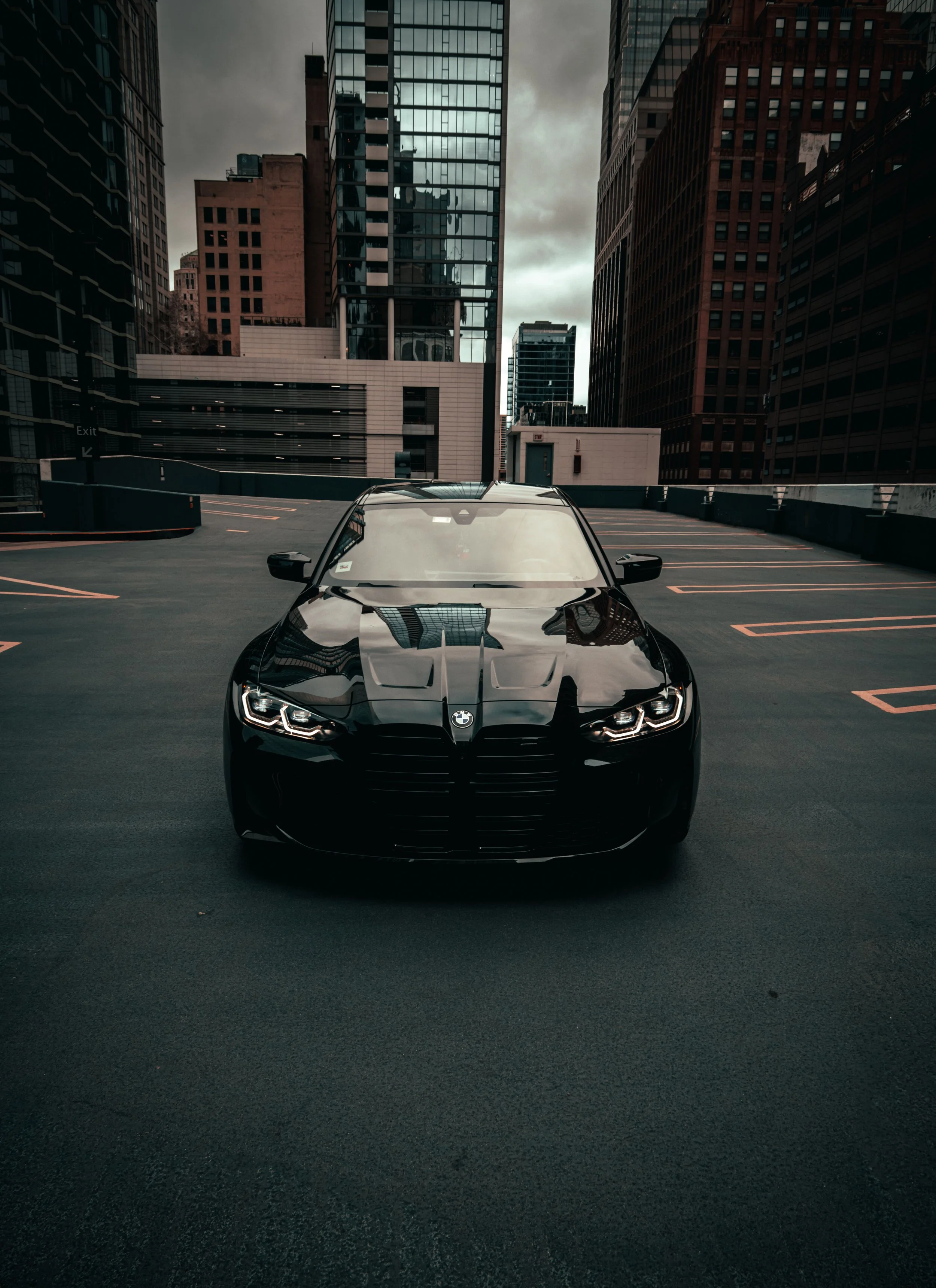 A black BMW car parked in a multi-level parking garage with tall city skyscrapers and cloudy sky in the background.
