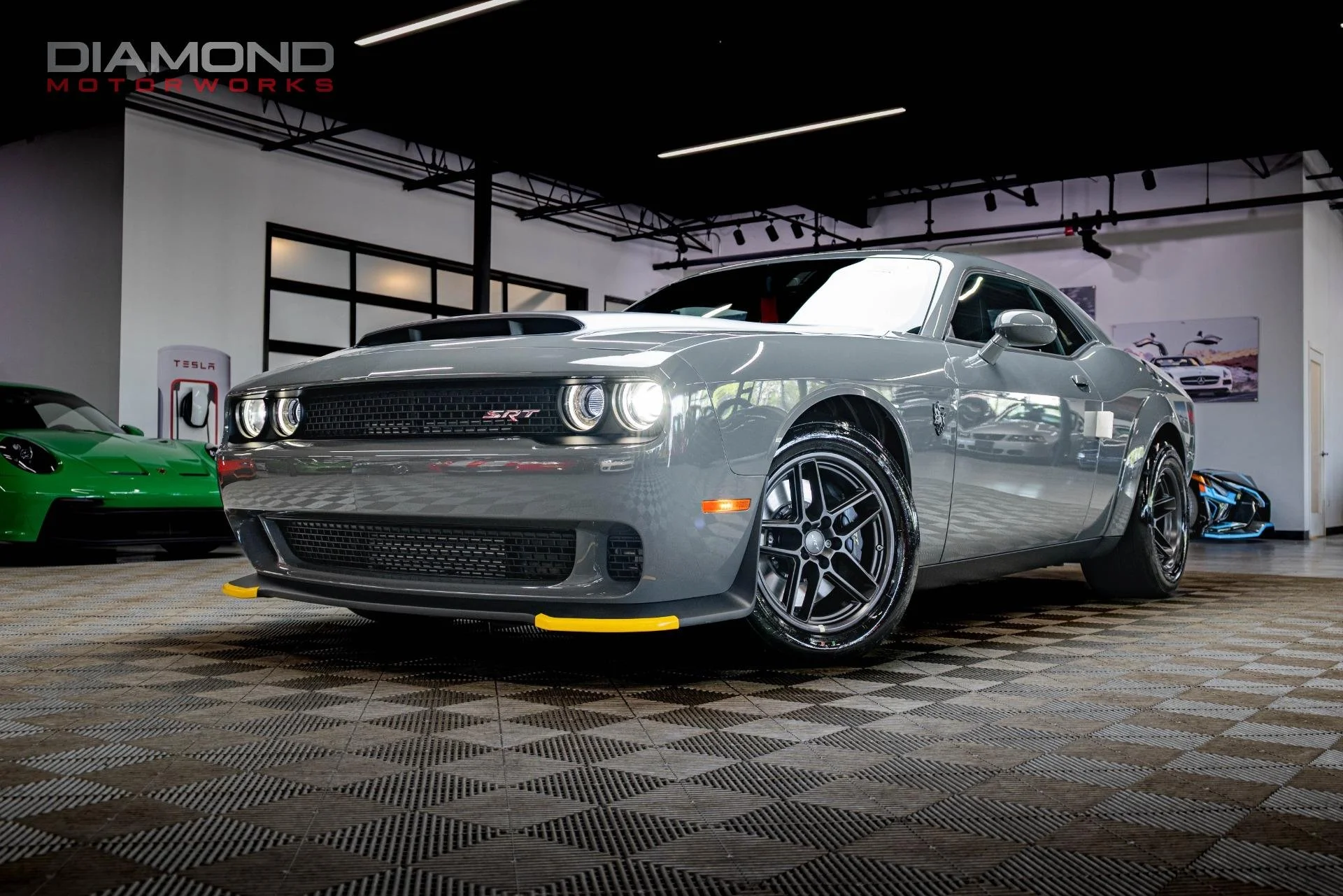 A silver muscle car parked inside a modern showroom with a black ceiling, black and white patterned floor, and a Tesla charging station visible in the background.