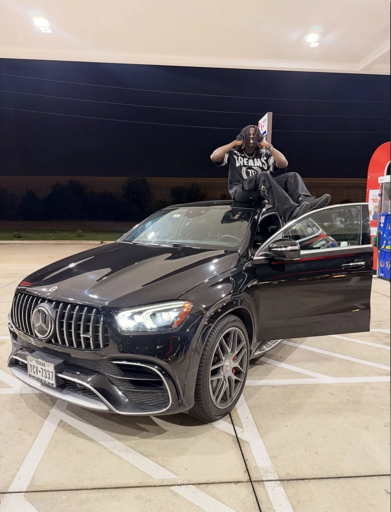A man with dreadlocks sitting on top of a black Mercedes-Benz SUV at a gas station during nighttime. The man is holding his headphones, and the vehicle's door is open. The gas station has a red and blue vending machine on the right side.