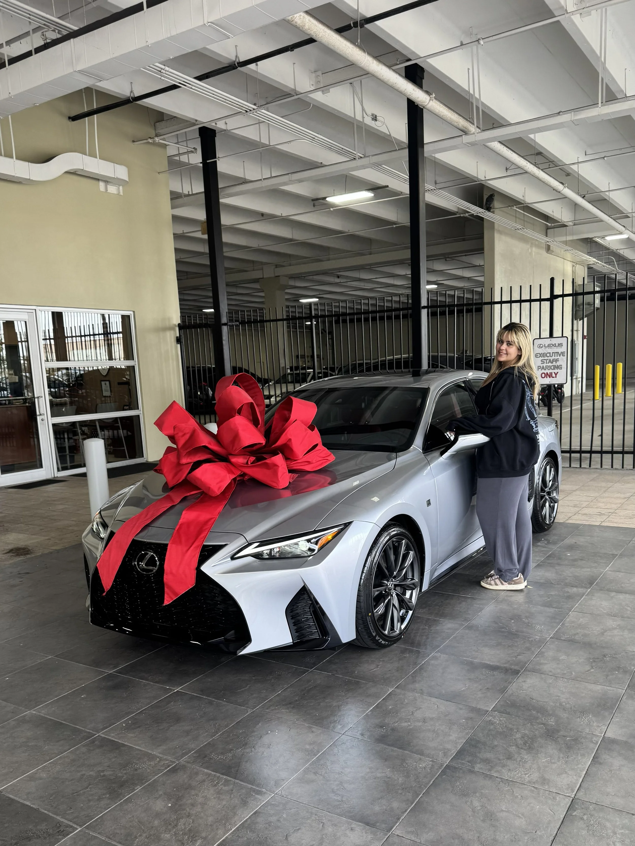 A woman stands next to a silver car decorated with a large red ribbon bow in an indoor parking garage. The car is new and shiny. The garage has a high ceiling with exposed pipes and black metal fencing. There is a sign that reads 'Executive Staff Parking Only' near the fence. The woman is smiling and wearing a black hoodie and gray sweatpants.