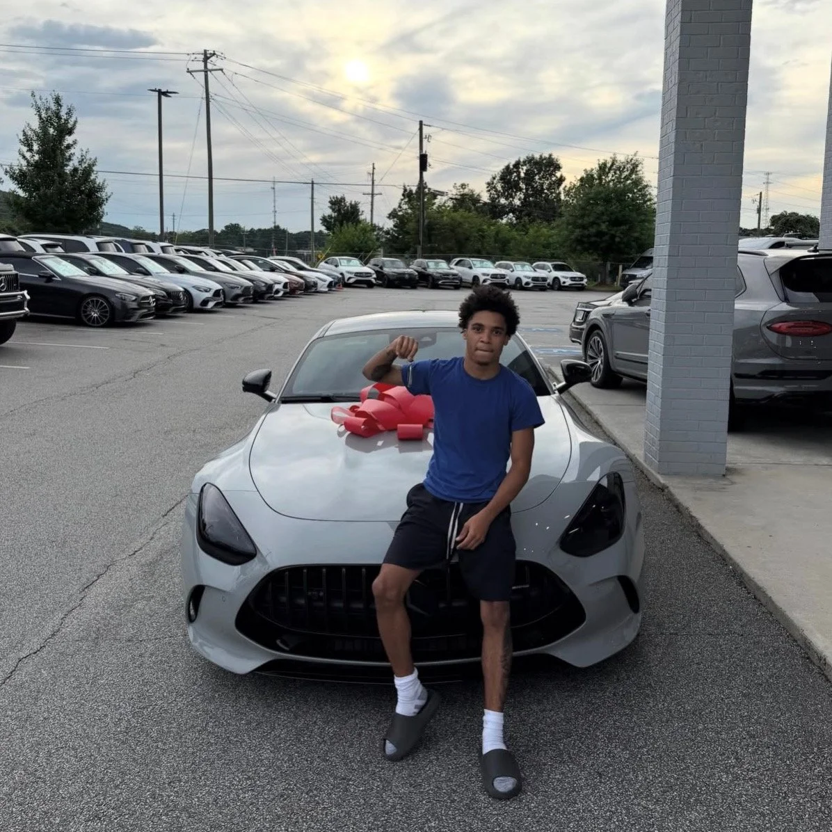 Young man standing next to a silver sports car with a large red bow on the hood at a car dealership lot.