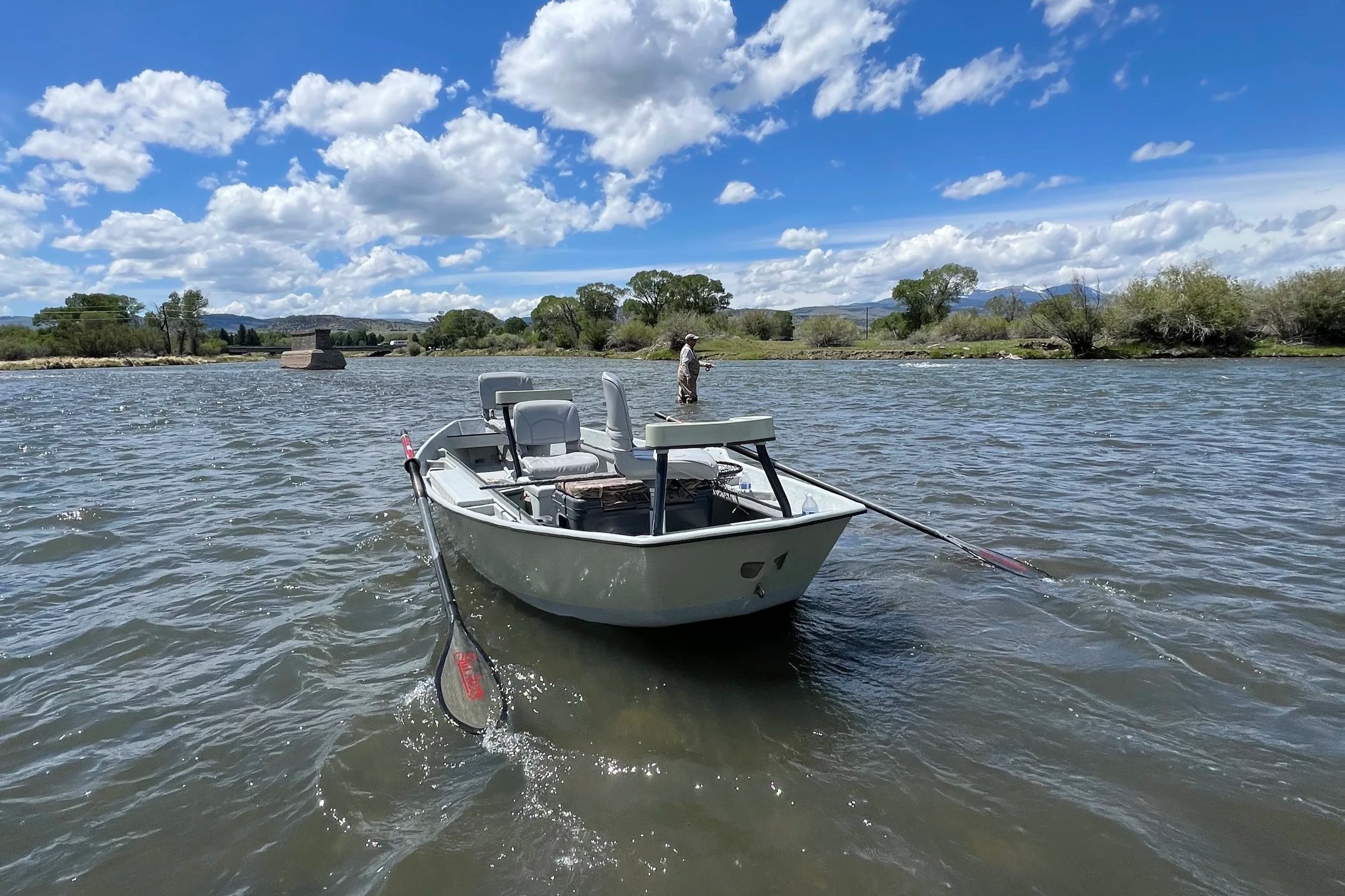 A person fishing in a river from a small boat on a sunny day with a blue sky and scattered clouds, lush greenery along the riverbanks, and distant mountains.