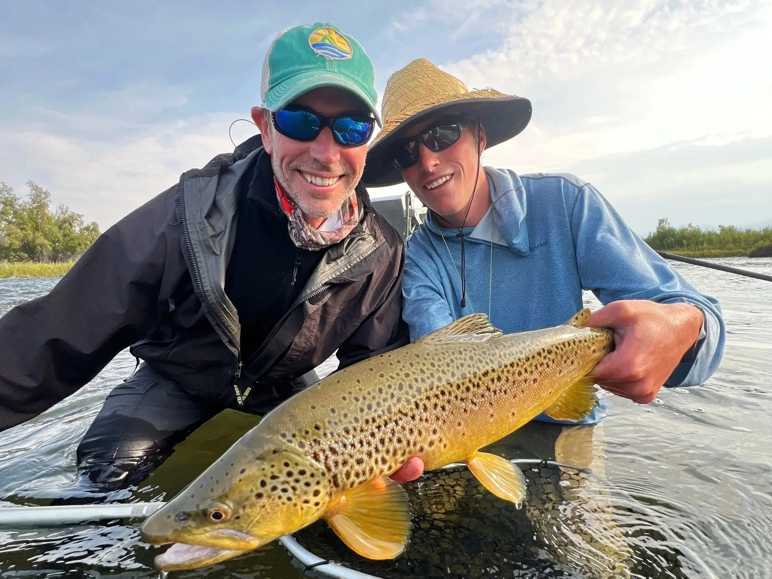 Two men smiling and posing with a large fish they caught, standing in a river with trees and a cloudy sky in the background.