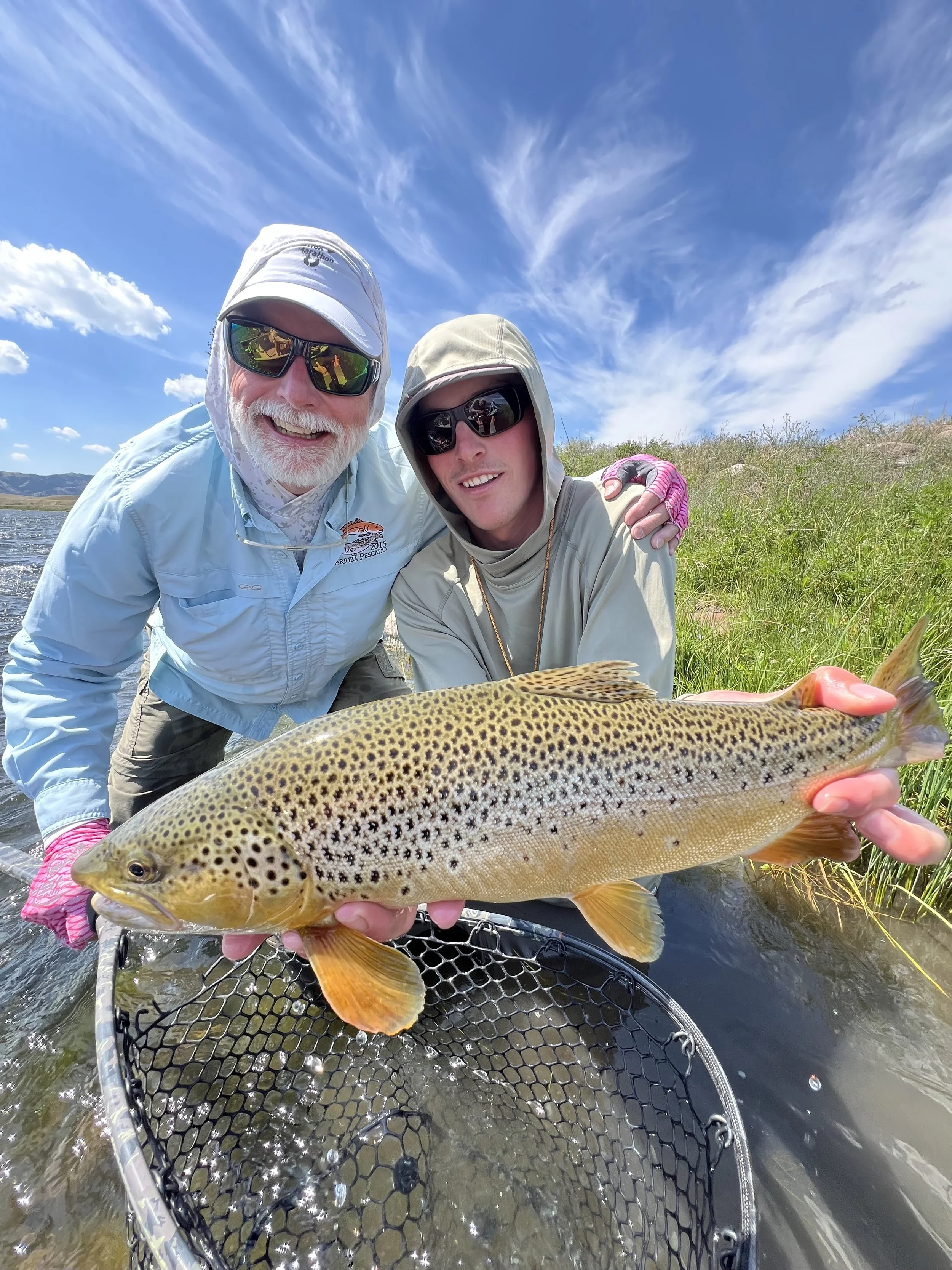 Two men are smiling and posing for a photo while holding a large, spotted fish with a pinkish tail, in a river with a grassy bank and blue sky with white clouds in the background.