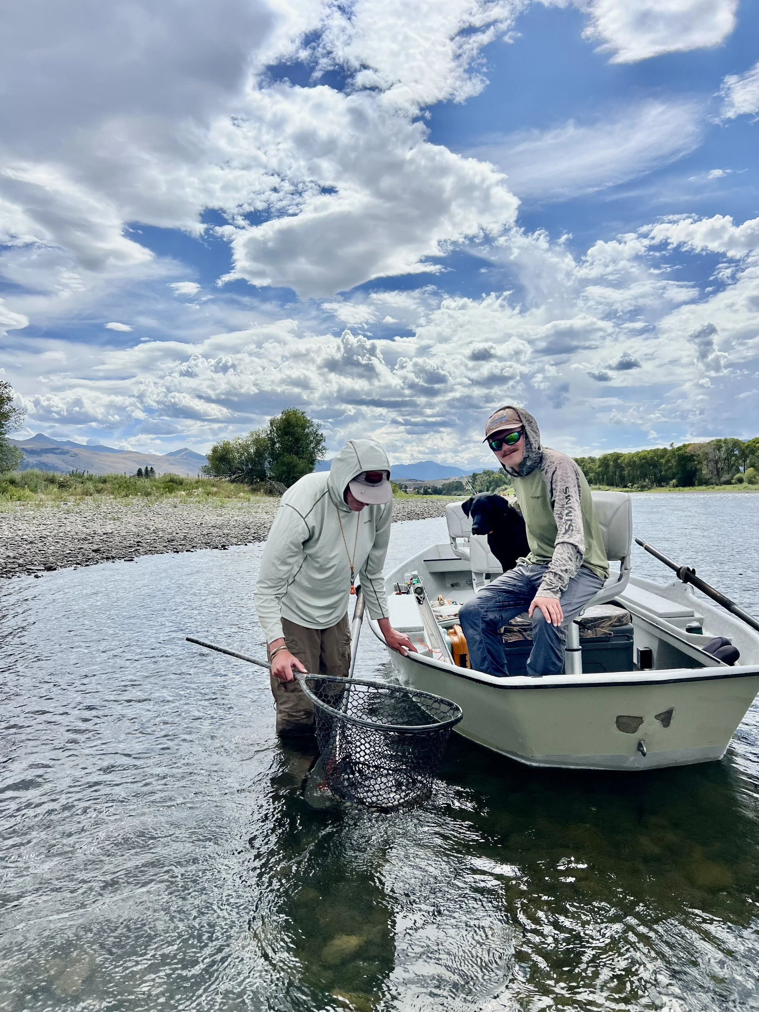 Two men and a black dog on a fishing boat in a river, with a rocky shore, trees, hills, and mountains in the background, under a partly cloudy sky.