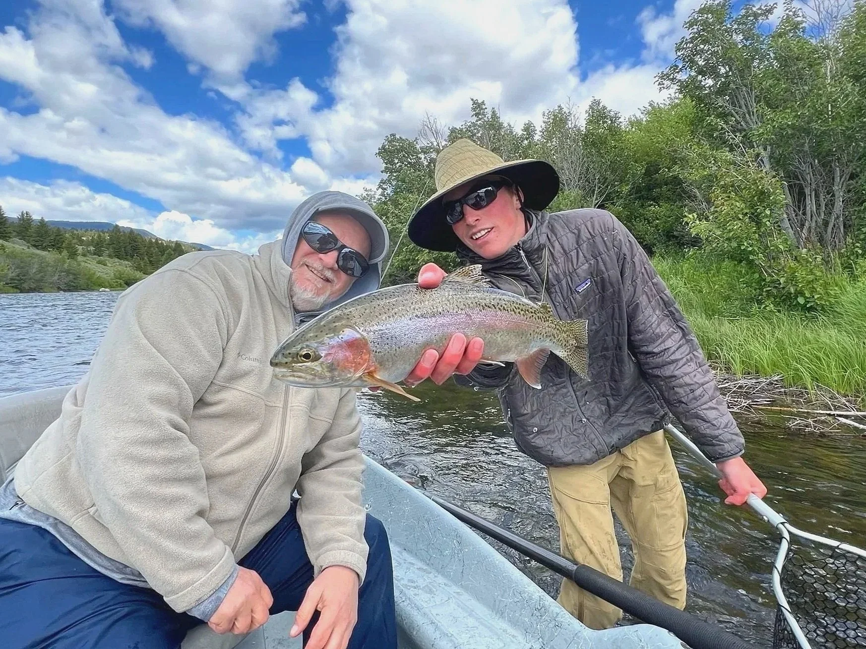 Two men in a boat holding a large fish they caught, one wearing a gray hoodie and sunglasses, the other in a wide-brimmed hat and jacket. They are outdoors near water and surrounded by trees and greenery.